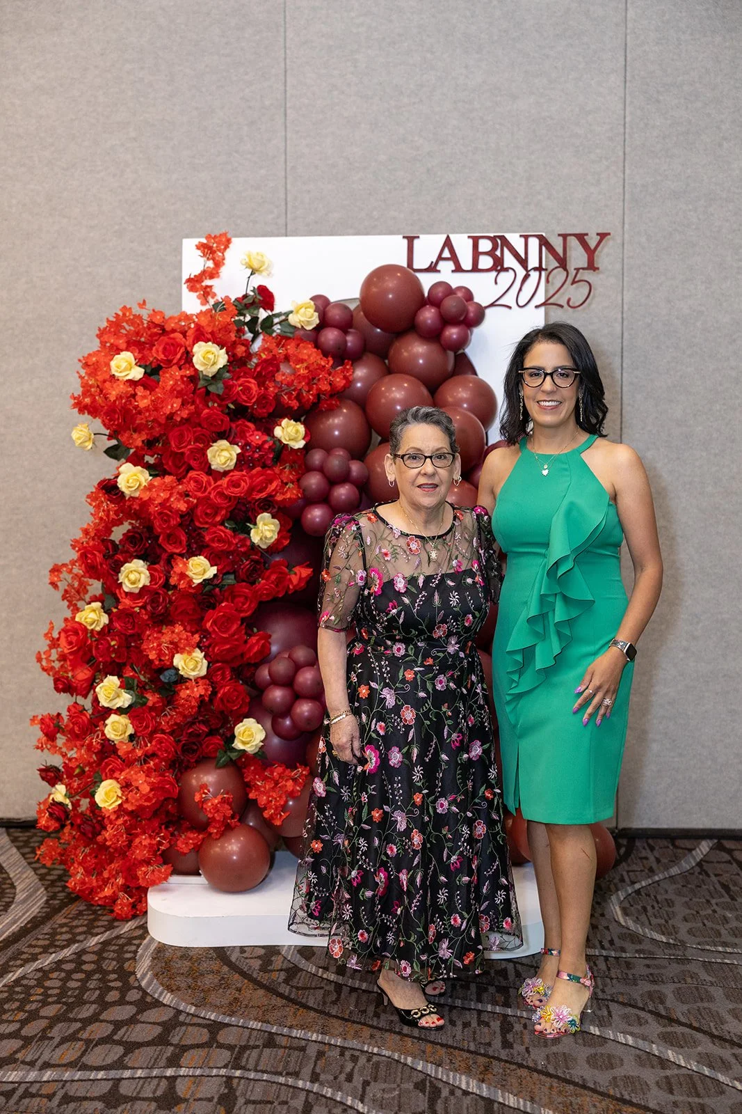 Two women standing in front of a floral and balloon display at an event, with a sign reading 'LABNYY 2025' in the background.