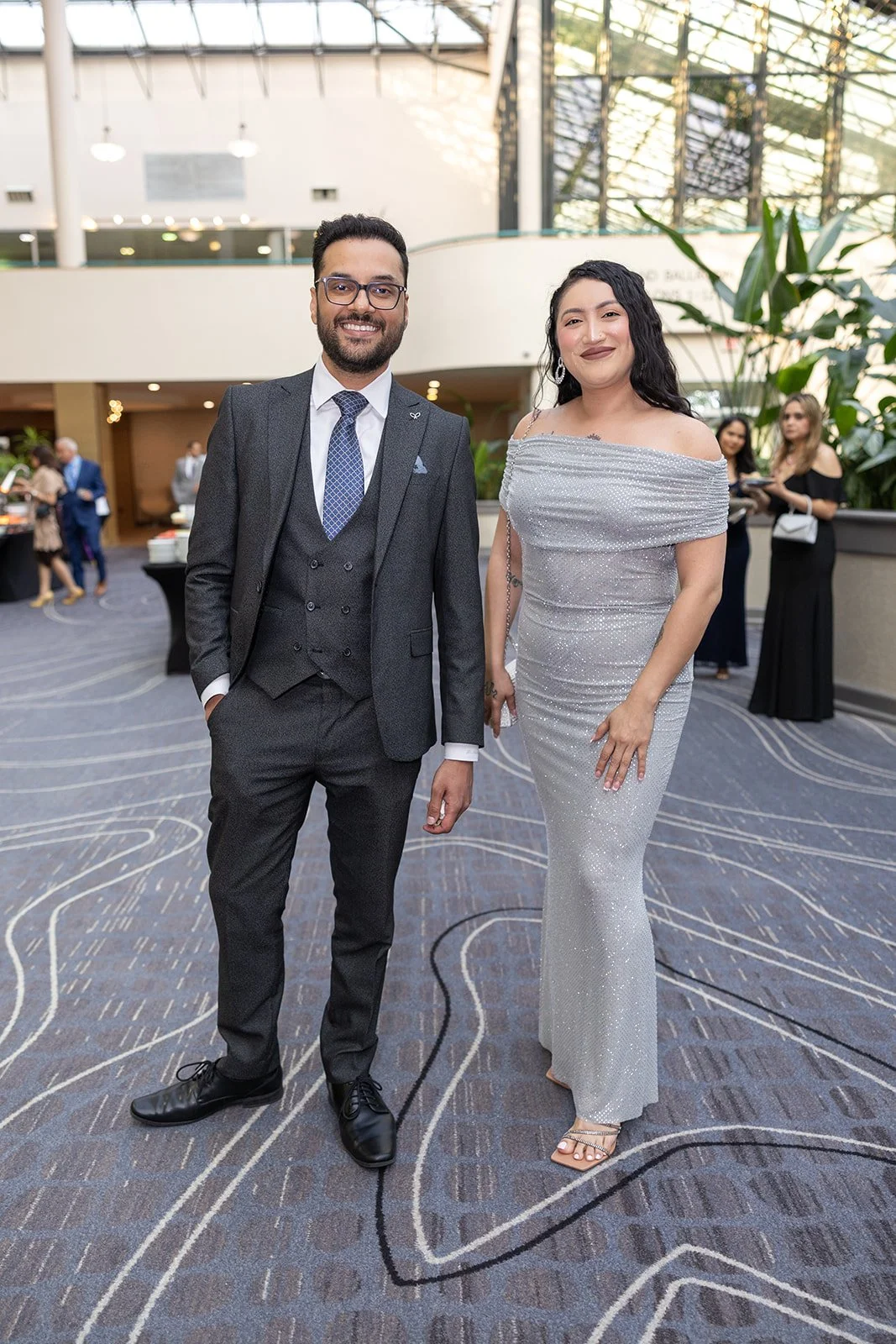 Man in a dark suit and woman in a silver dress standing and smiling at an indoor event.