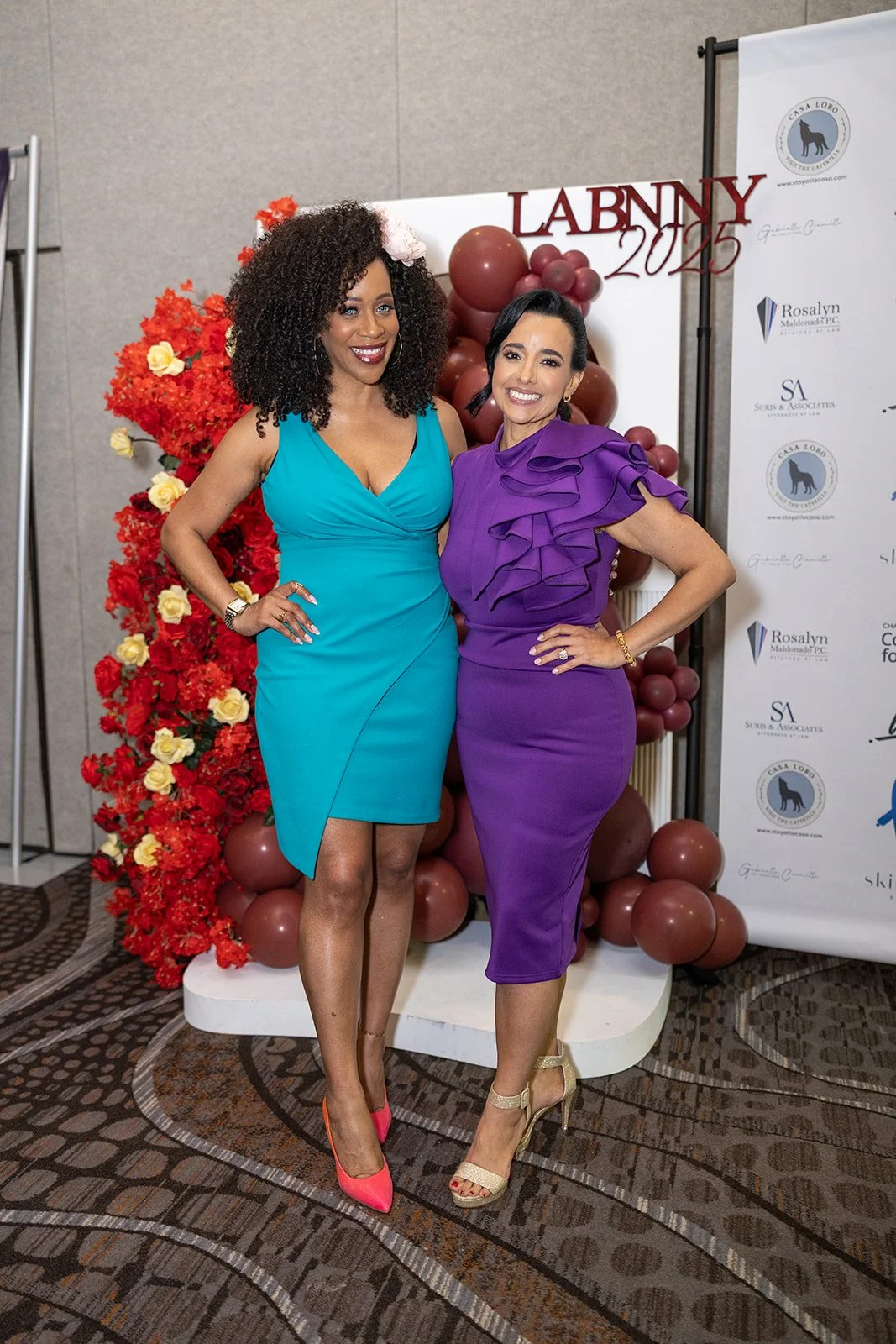 Two women dressed in colorful dresses standing in front of a backdrop with balloons and flowers, smiling for the camera at an event called LABNNY 2025.