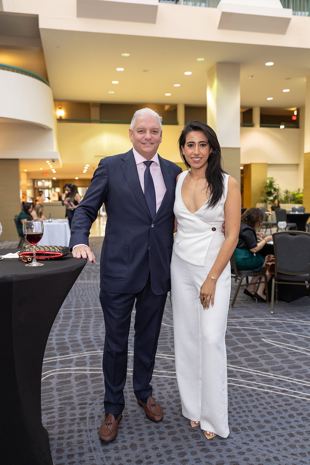 A man and woman stand together in a hotel lobby, smiling. The man is wearing a dark blue suit with a tie, and the woman is in a white dress. There are tables and other guests in the background.