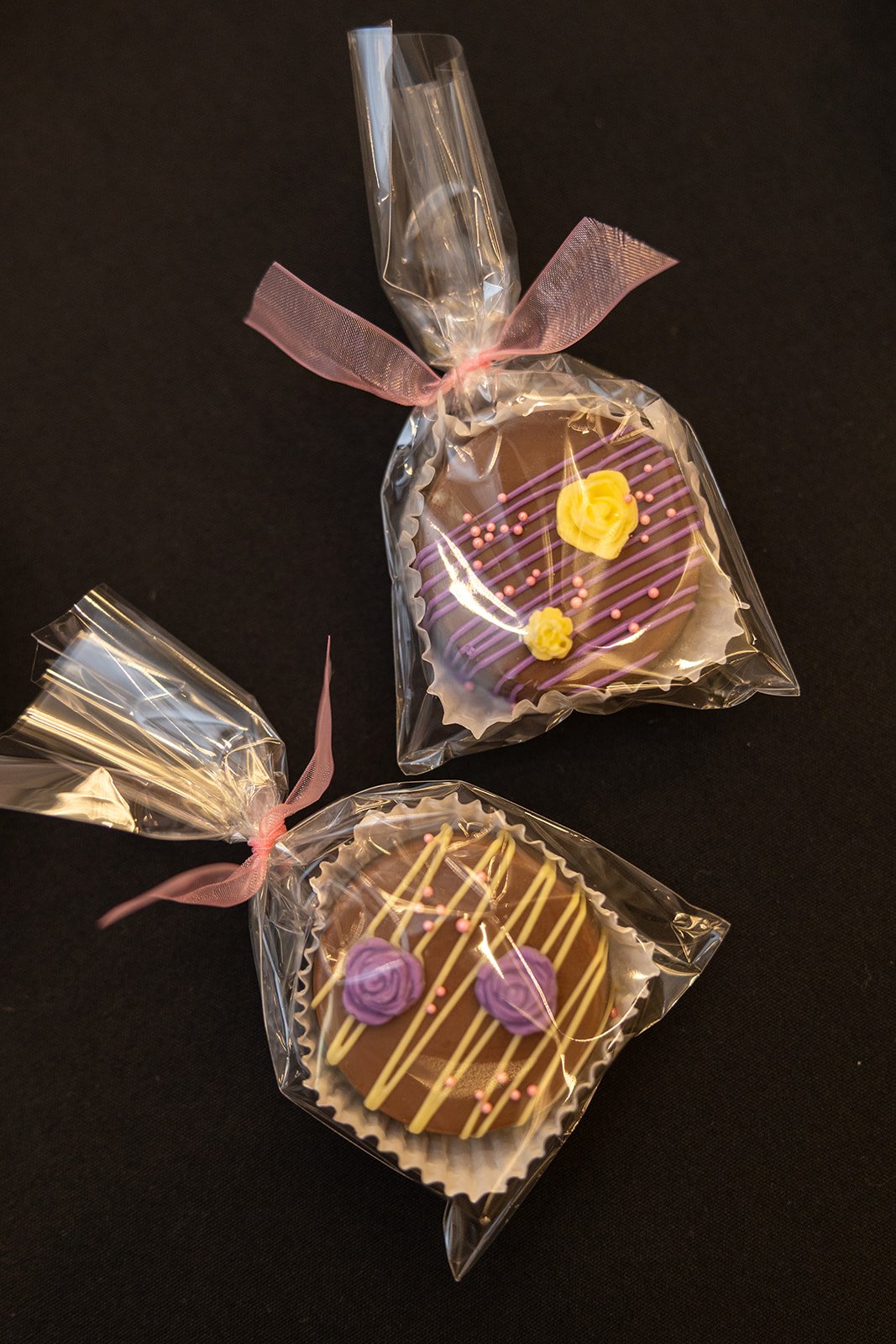 Two decorated chocolate candies wrapped in clear plastic with pink ribbons, placed on a black surface.
