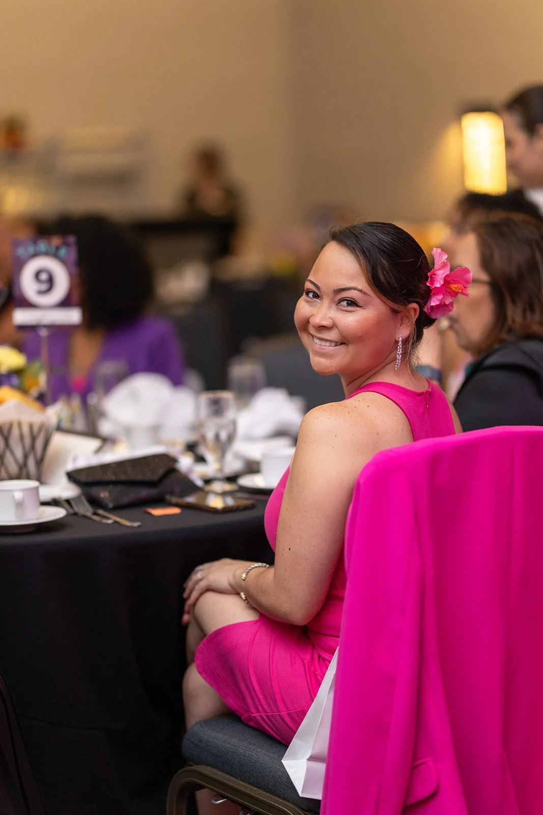 A woman in a pink dress with a pink flower in her hair smiling at a formal event.