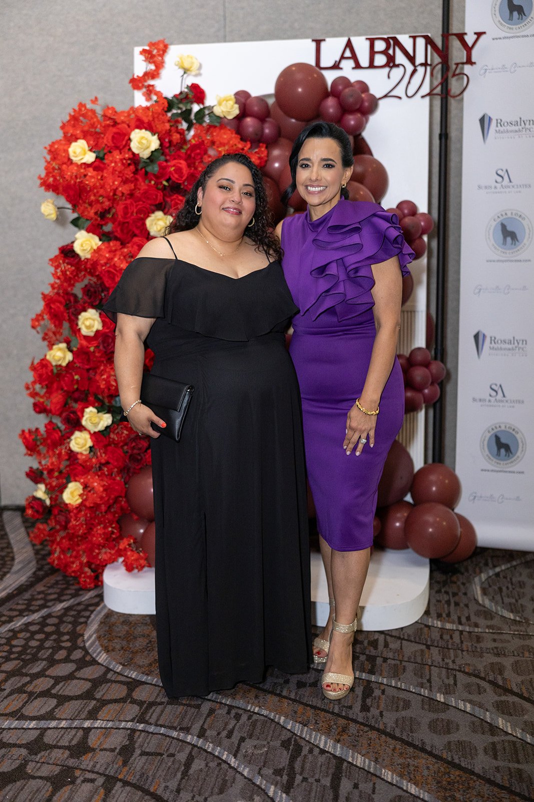 Two women in formal attire standing in front of a decorative backdrop with red and yellow flowers and balloons, at an event called 'LABNMY 2025'.