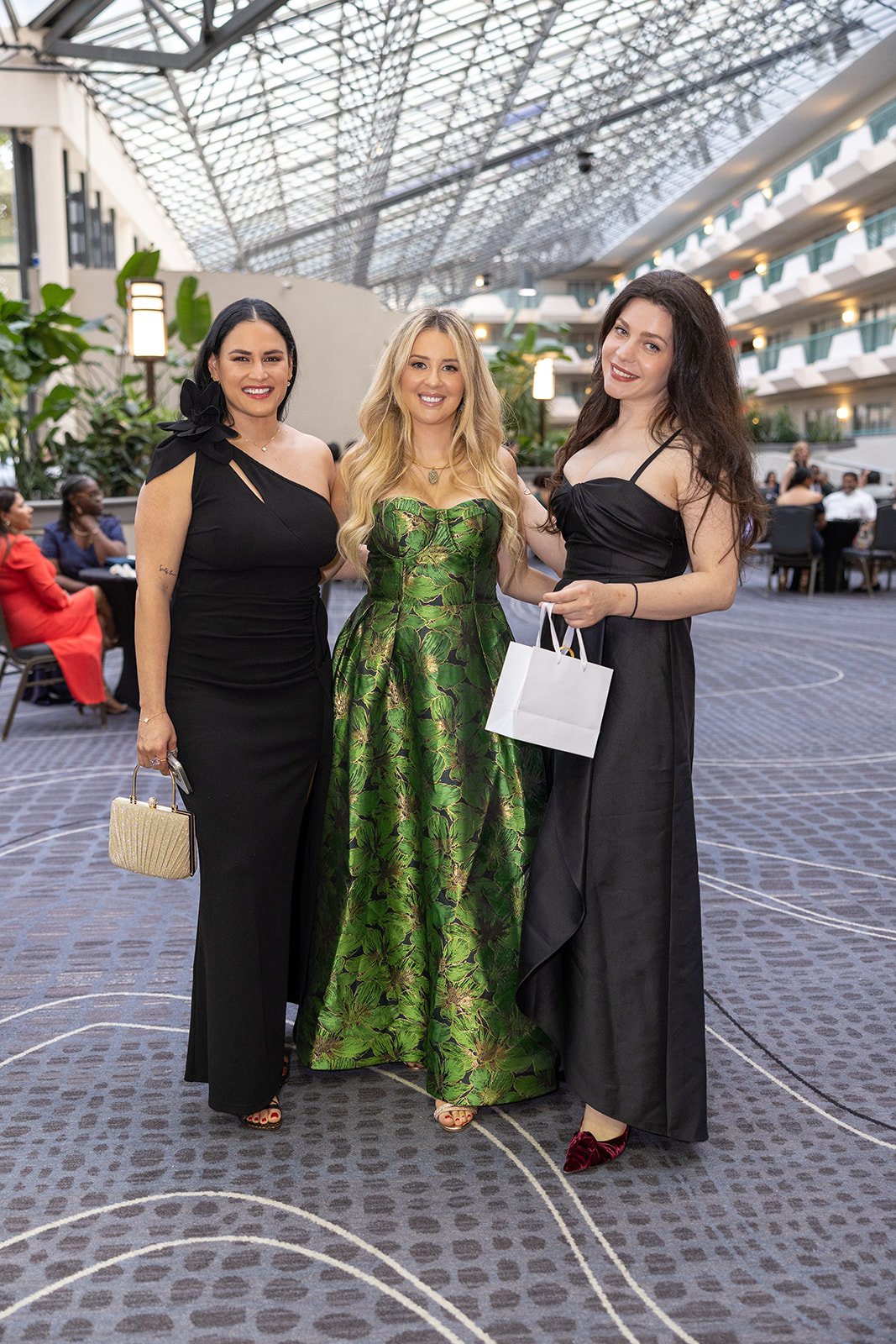 Three women in evening dress standing together inside a large atrium, smiling at the camera. The woman on the left wears a black dress, the woman in the middle wears a green patterned gown, and the woman on the right wears a black dress and holds a w