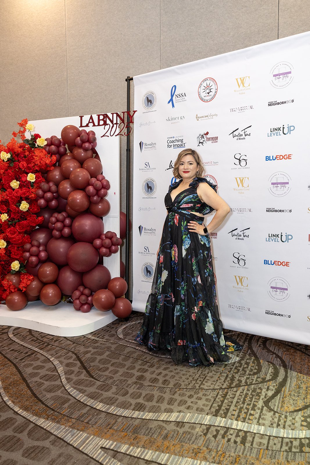 A woman in a black floral dress standing with hand on hip at a formal event with a decorated backdrop and a balloon display.