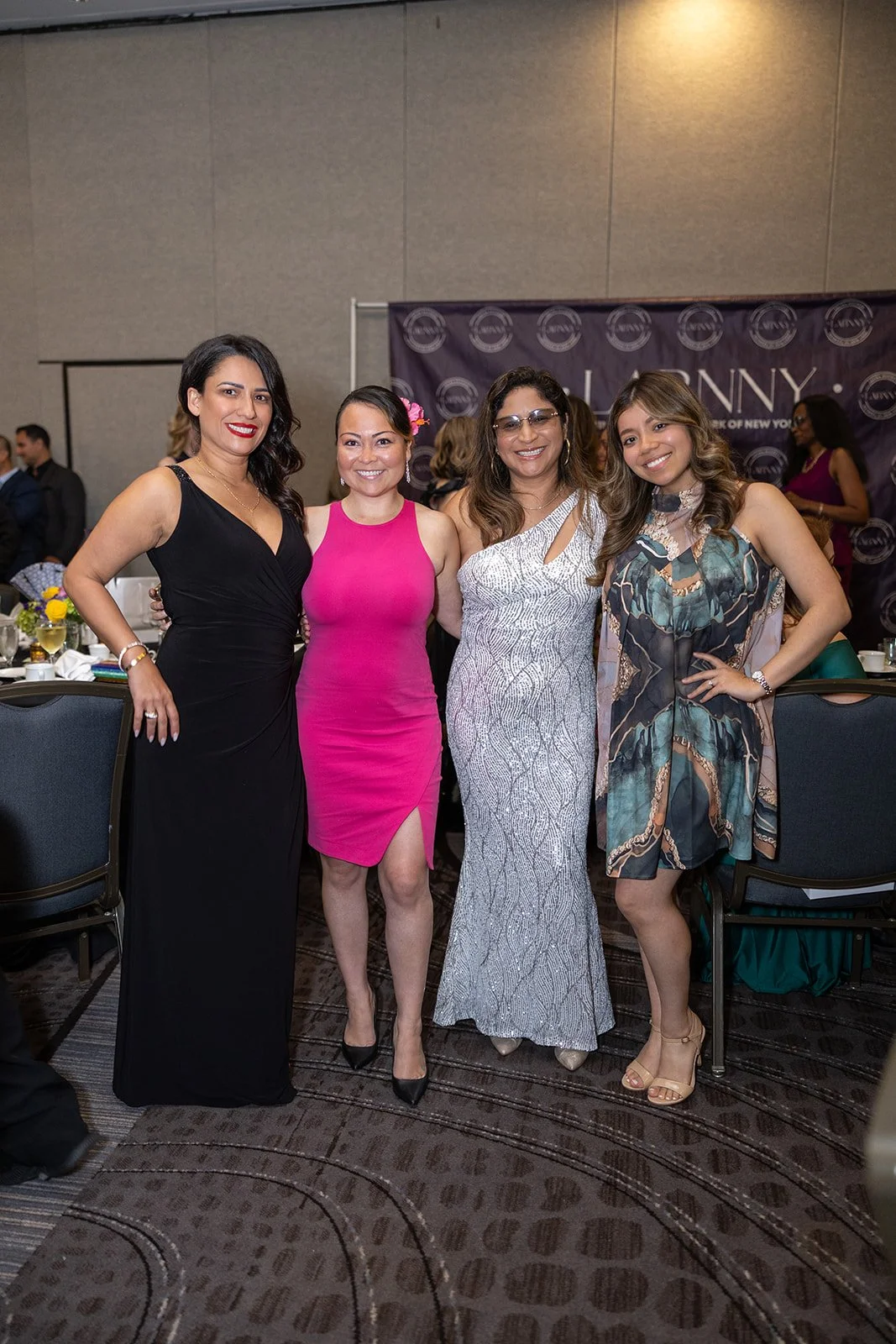 Four women in formal dresses standing together at an indoor event with tables and a backdrop behind them.