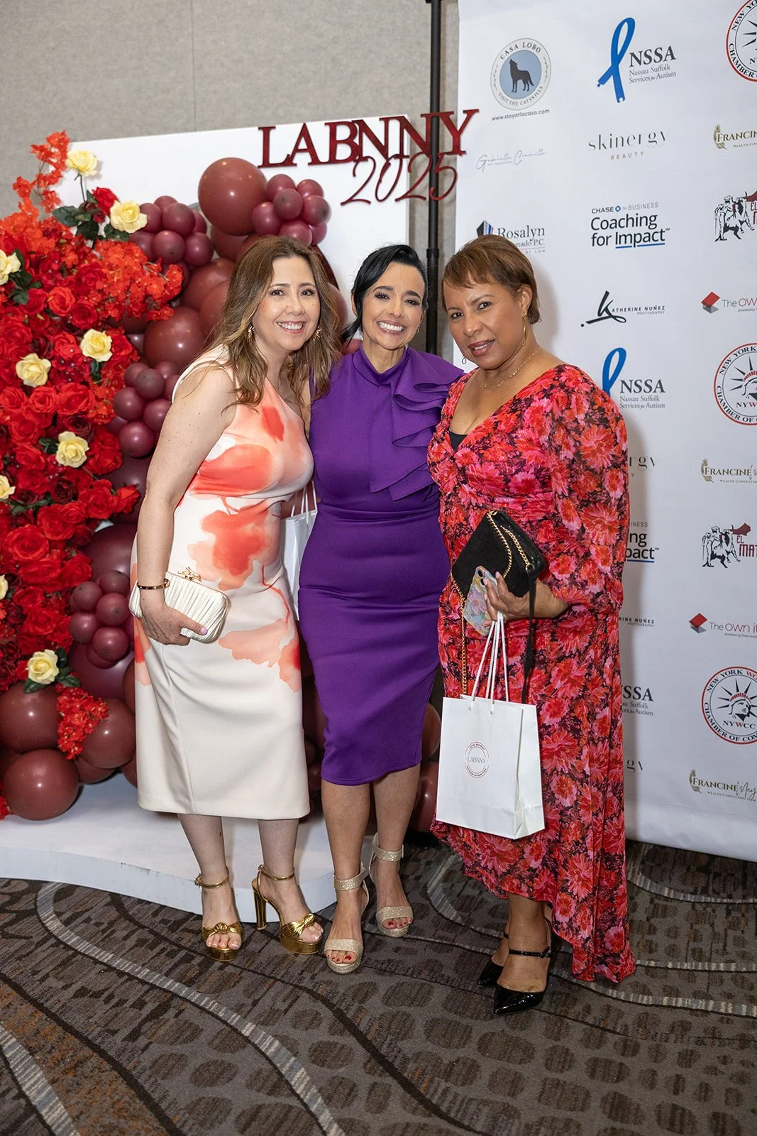 Three women in colorful dresses standing together at the LABNNY 2023 event, with a backdrop featuring logos and a decorative arrangement of red, pink, and yellow flowers and balloons.