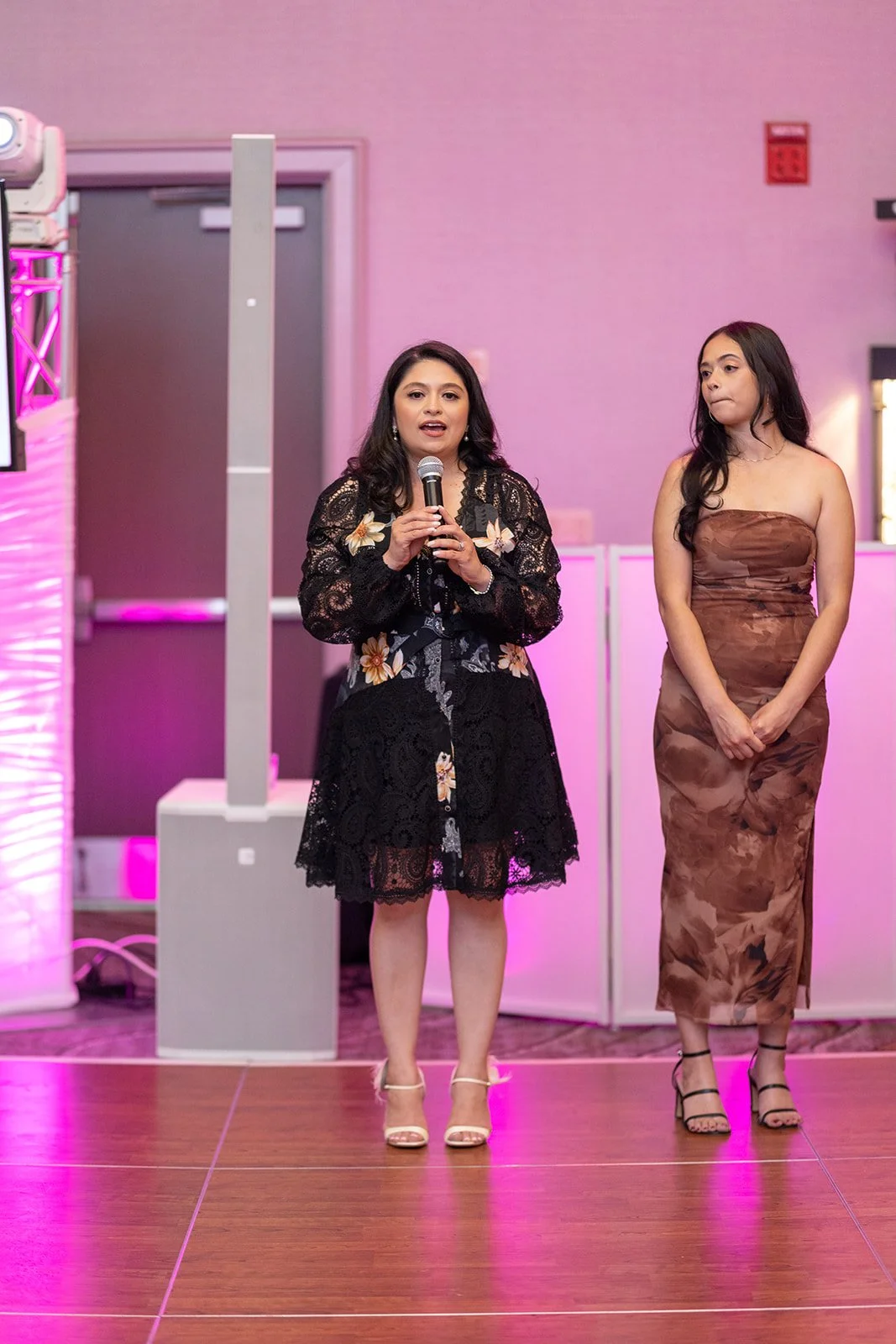 Two women standing on a stage with pink lighting, one speaking into a microphone. One woman is wearing a black lace dress with floral embroidery, and the other is in a strapless brown dress with a marbled pattern.