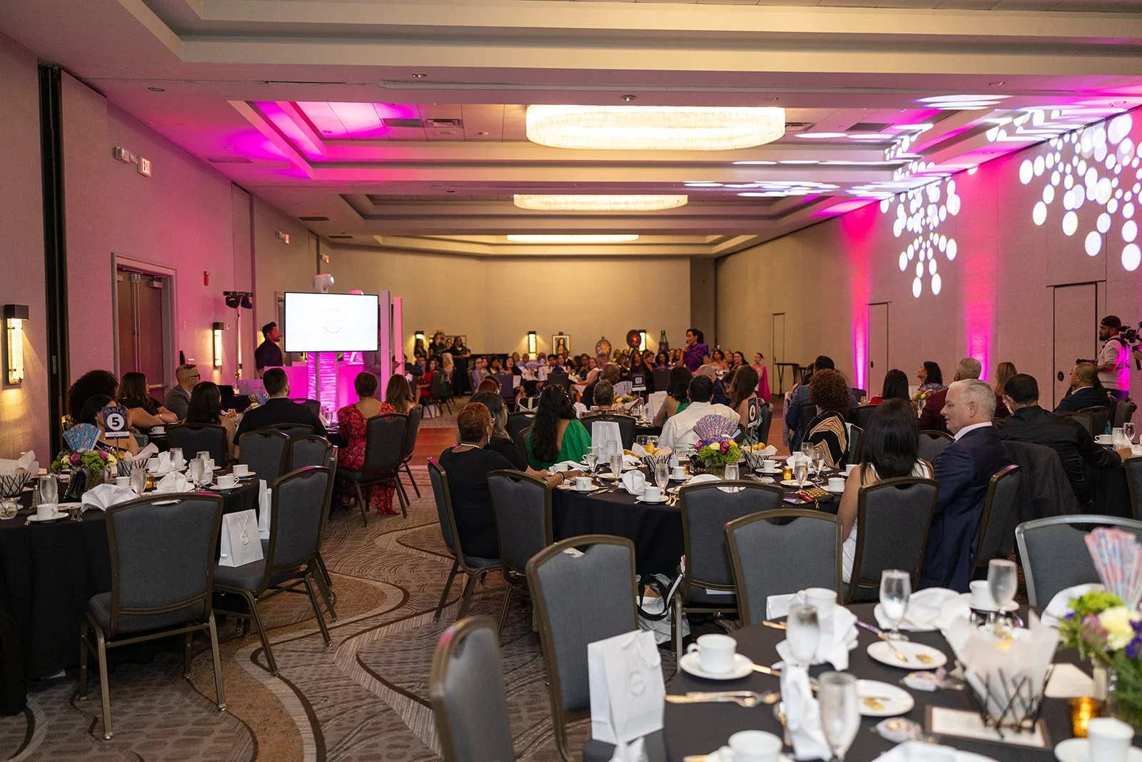 A large banquet hall filled with round tables set for a formal event, with people seated and a stage at the front with purple lighting and a large screen.