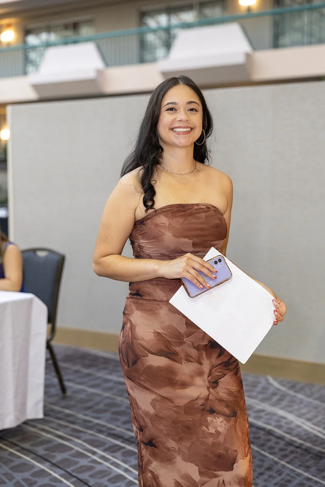 A woman in a strapless brown dress smiling, holding a smartphone, a piece of paper, and a pen in an indoor event space.