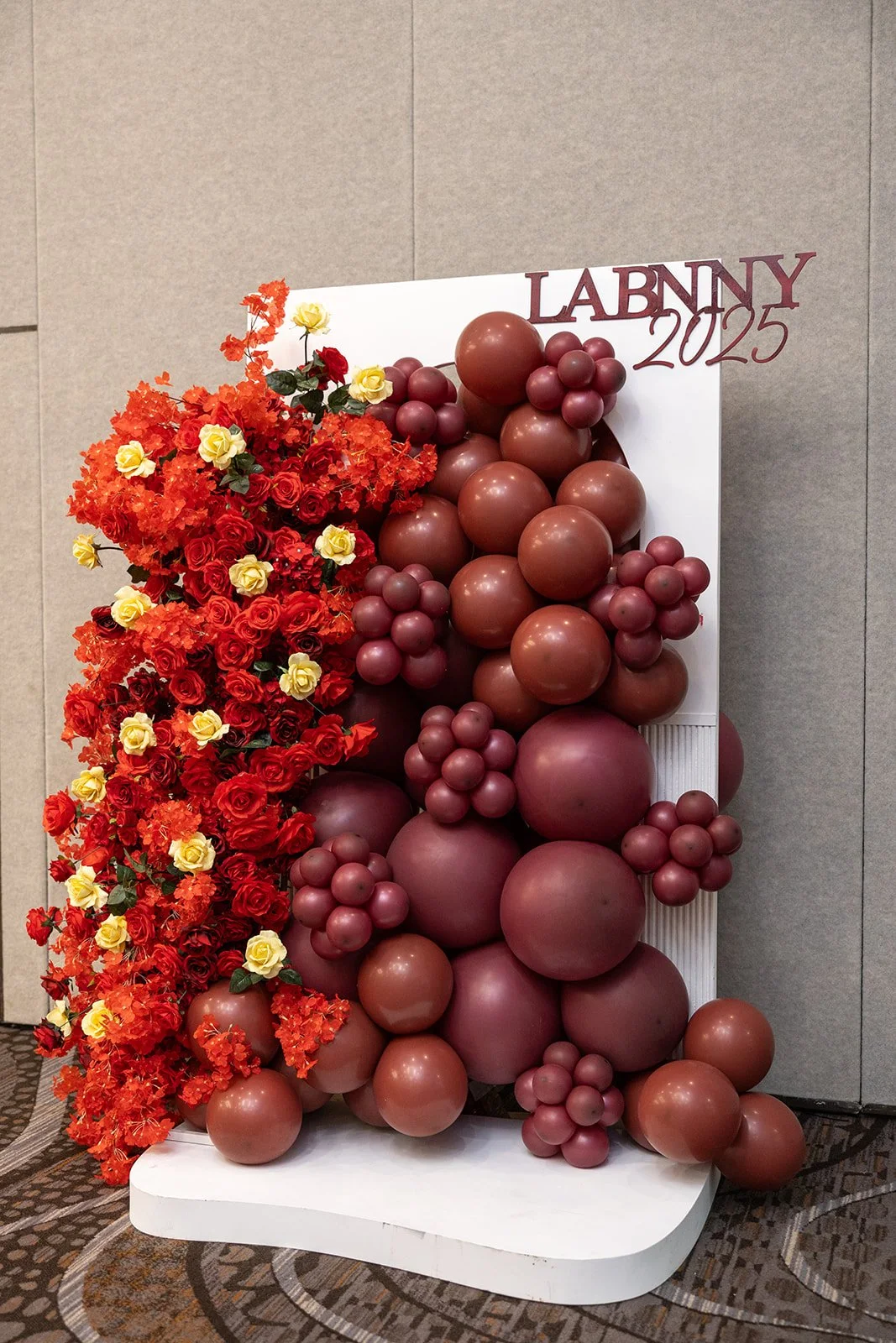 A decorative display with red and yellow flowers and a cluster of red and maroon balloons, with the words 'LABNY 2025' at the top.