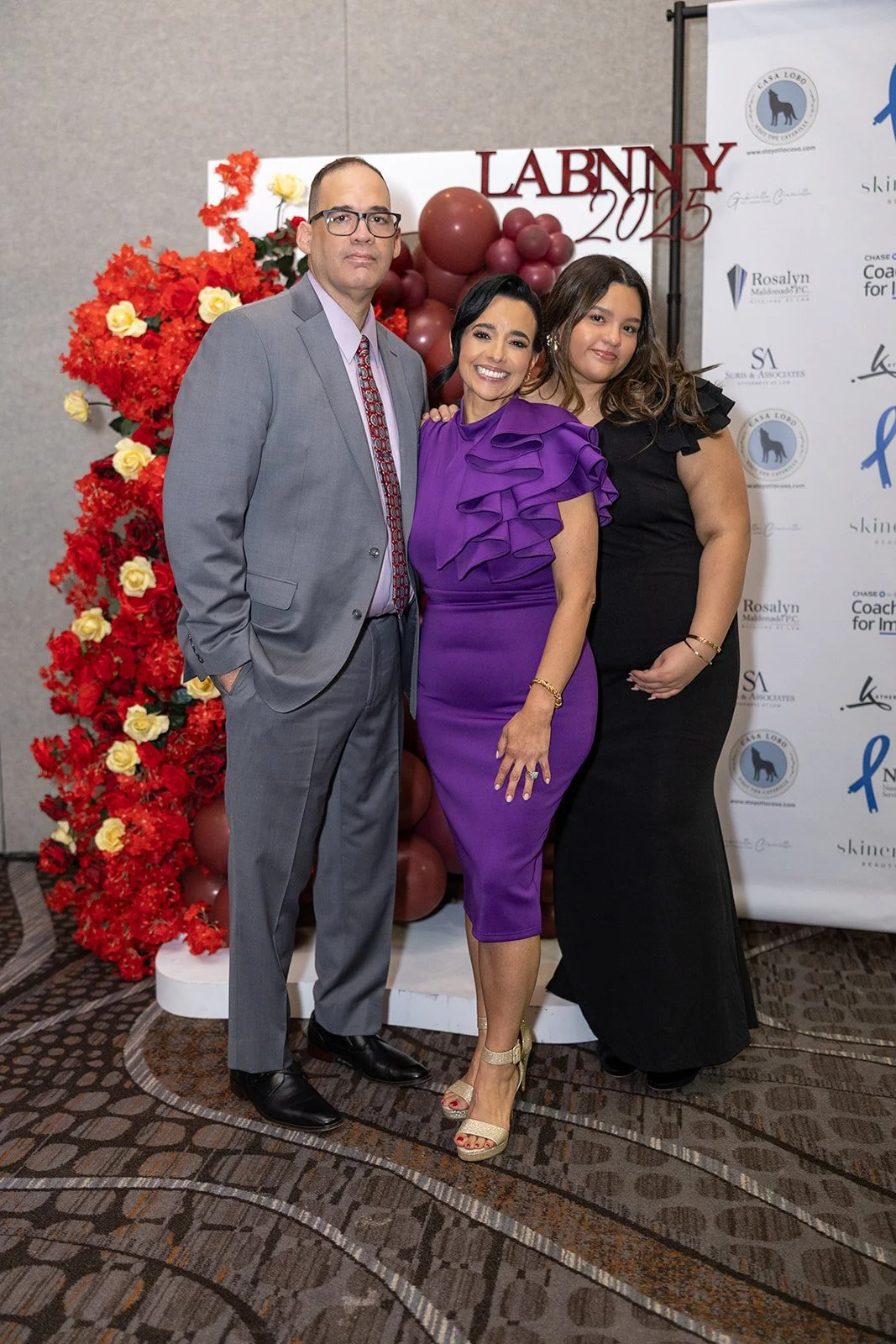 Three people dressed in formal attire posing at an event, with a backdrop featuring various logos and decorations including balloons and flowers, and a sign reading "LABNNY 2025."