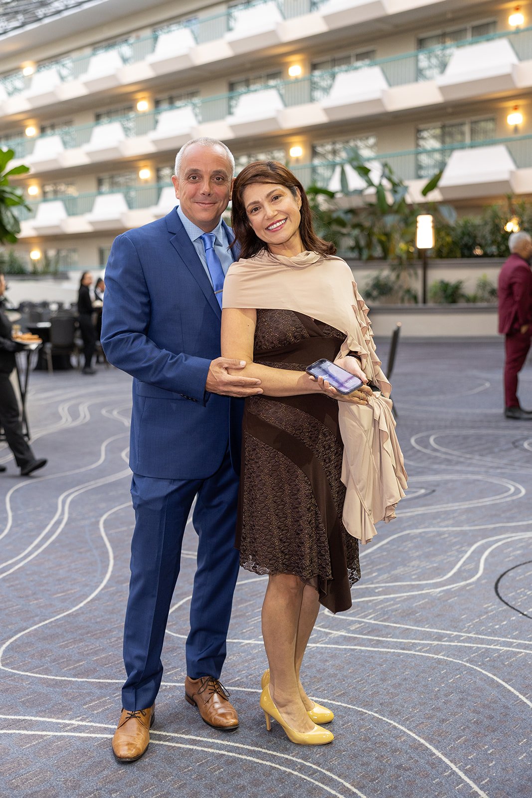 A man and woman standing together at an indoor event. The man is dressed in a blue suit and the woman is wearing a black and beige dress with yellow high heels. They are smiling and the woman is holding a phone. Background shows a decorated atrium wi
