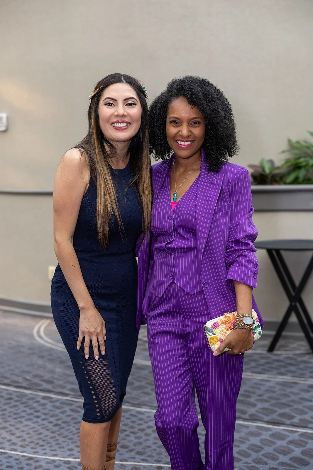 Two women standing together indoors, smiling at the camera. One wears a navy blue dress, and the other wears a purple pinstriped suit and holding a floral clutch.