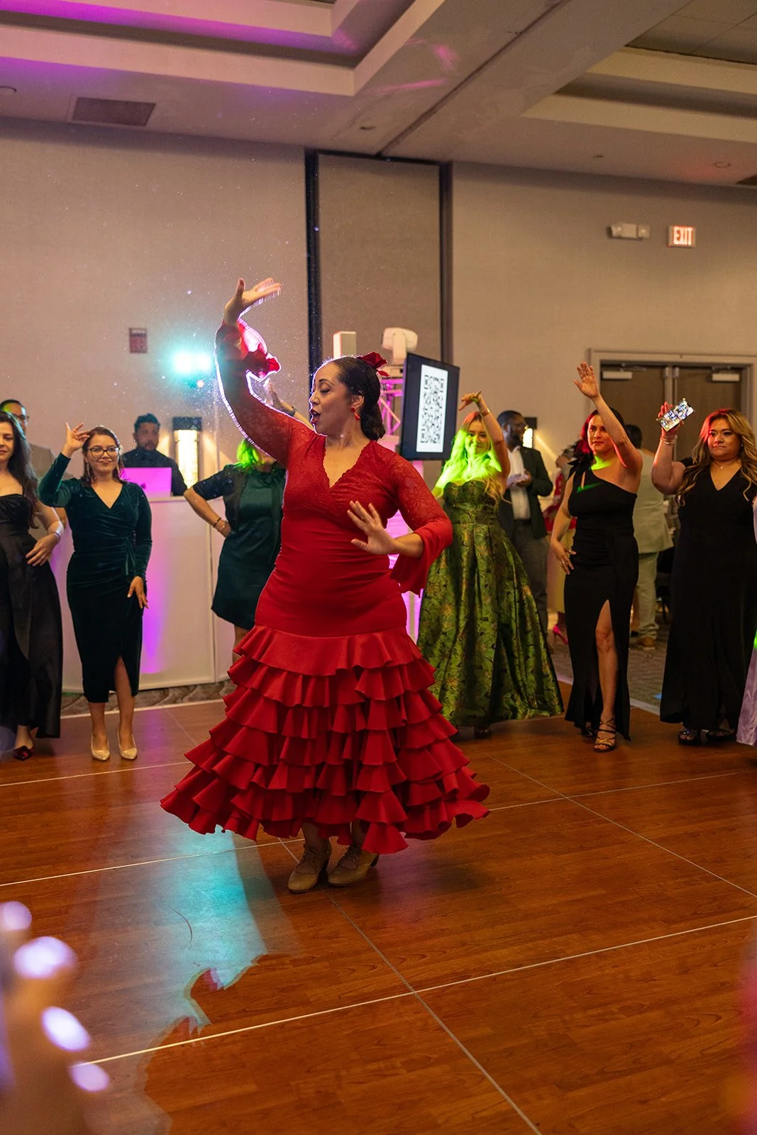 A woman in a red flamenco-style dress dancing on a wooden dance floor, surrounded by other people in formal attire at an indoor celebration or event.