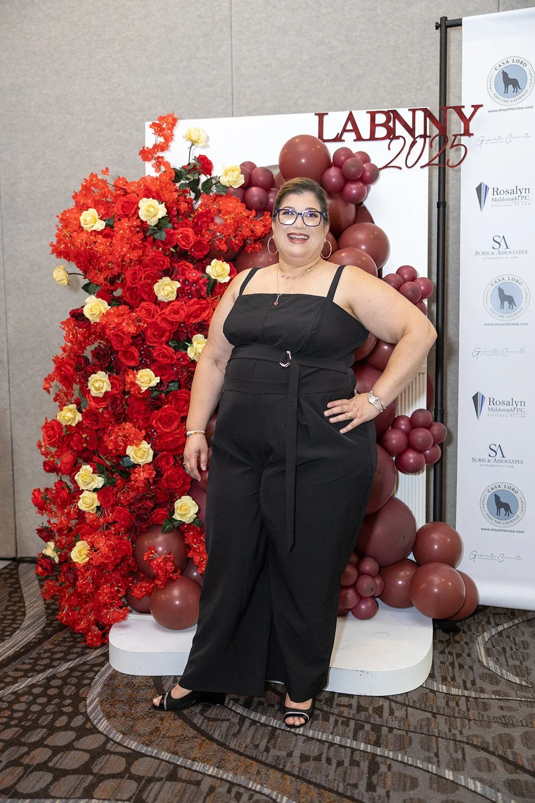 A woman in black outfit and glasses standing in front of red and yellow floral decorations with balloons and a banner for LABNY 2023.