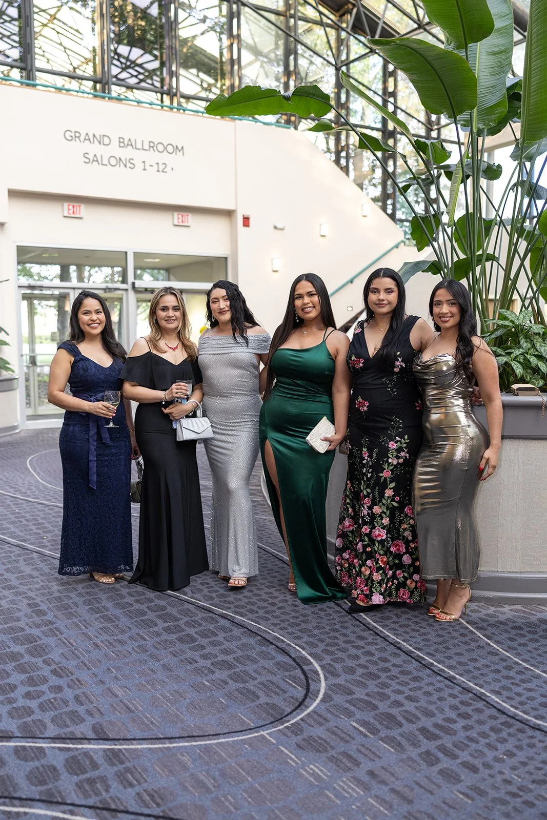 Six women dressed in formal evening gowns standing together at an indoor event, smiling at the camera. The background shows large plants, glass windows, and a sign indicating 'Grand Ballroom Salons 1-12'.