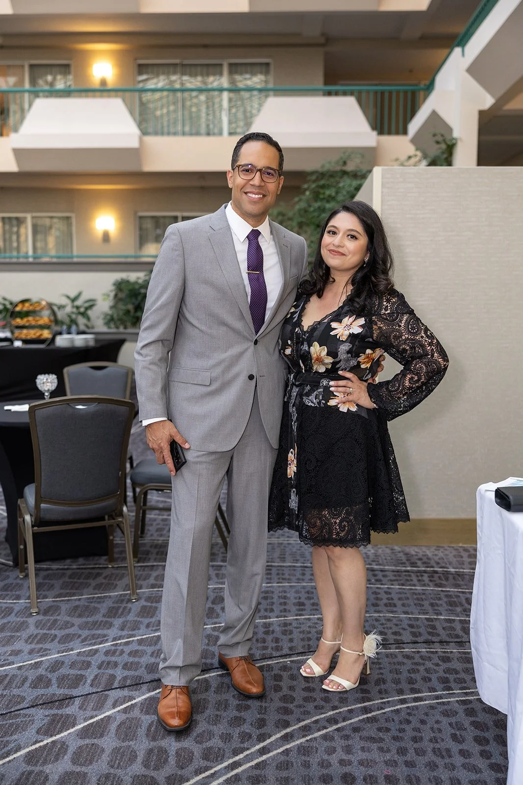 A man and woman posing together at a formal event in an indoor venue, with tables and chairs in the background.