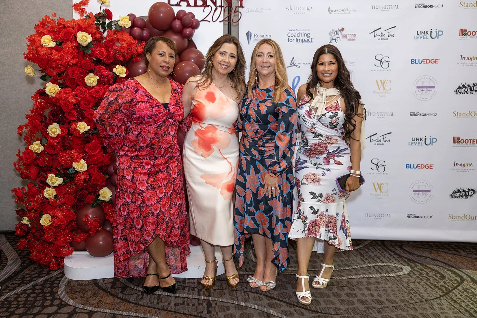 Four women standing together at an event, dressed in floral and colorful dresses, with a backdrop displaying event sponsors and decorations of red and yellow roses and balloons.