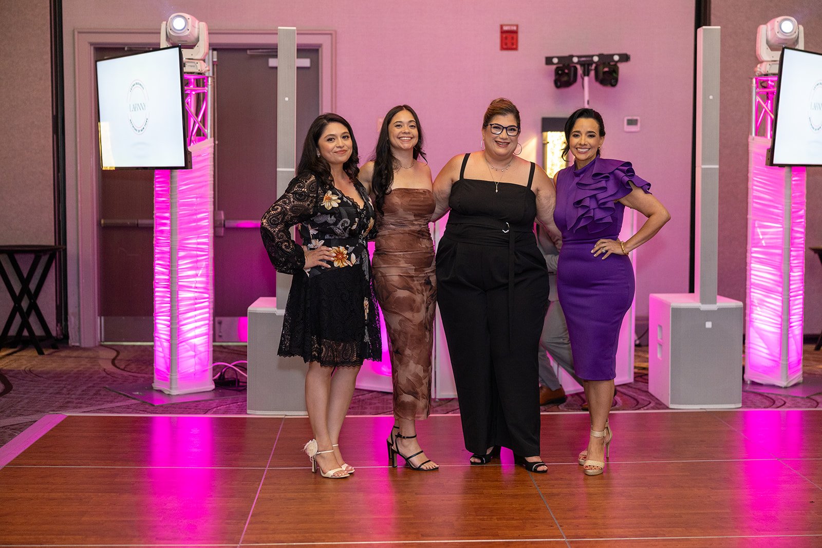 Four women standing together on a dance floor at an indoor event, under purple lighting with two large screens and DJ equipment in the background.