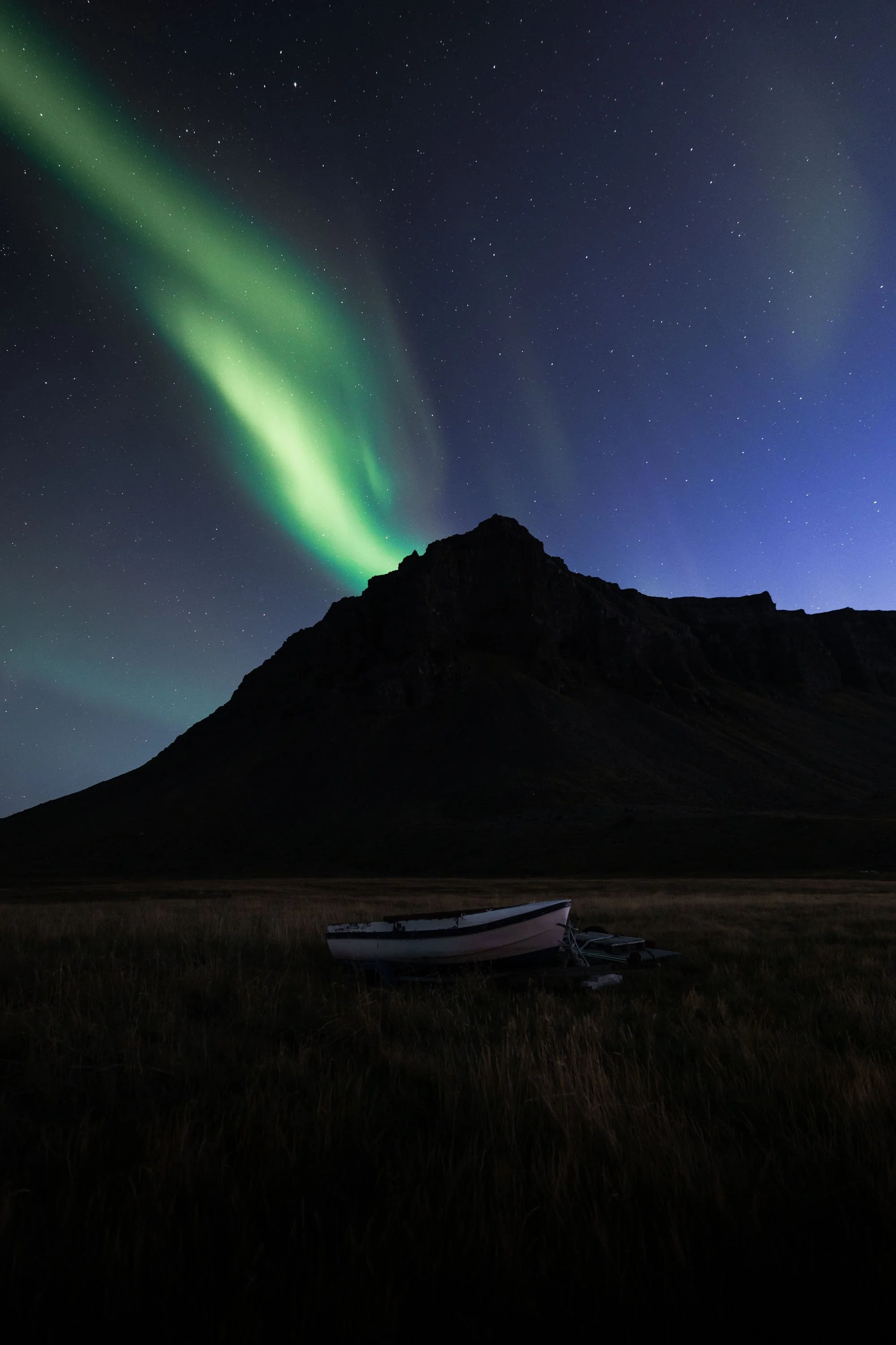 A vibrant green aurora borealis (Northern Lights) dancing in the night sky over a fjord with a boat in the foreground in the Westfjords, Iceland.