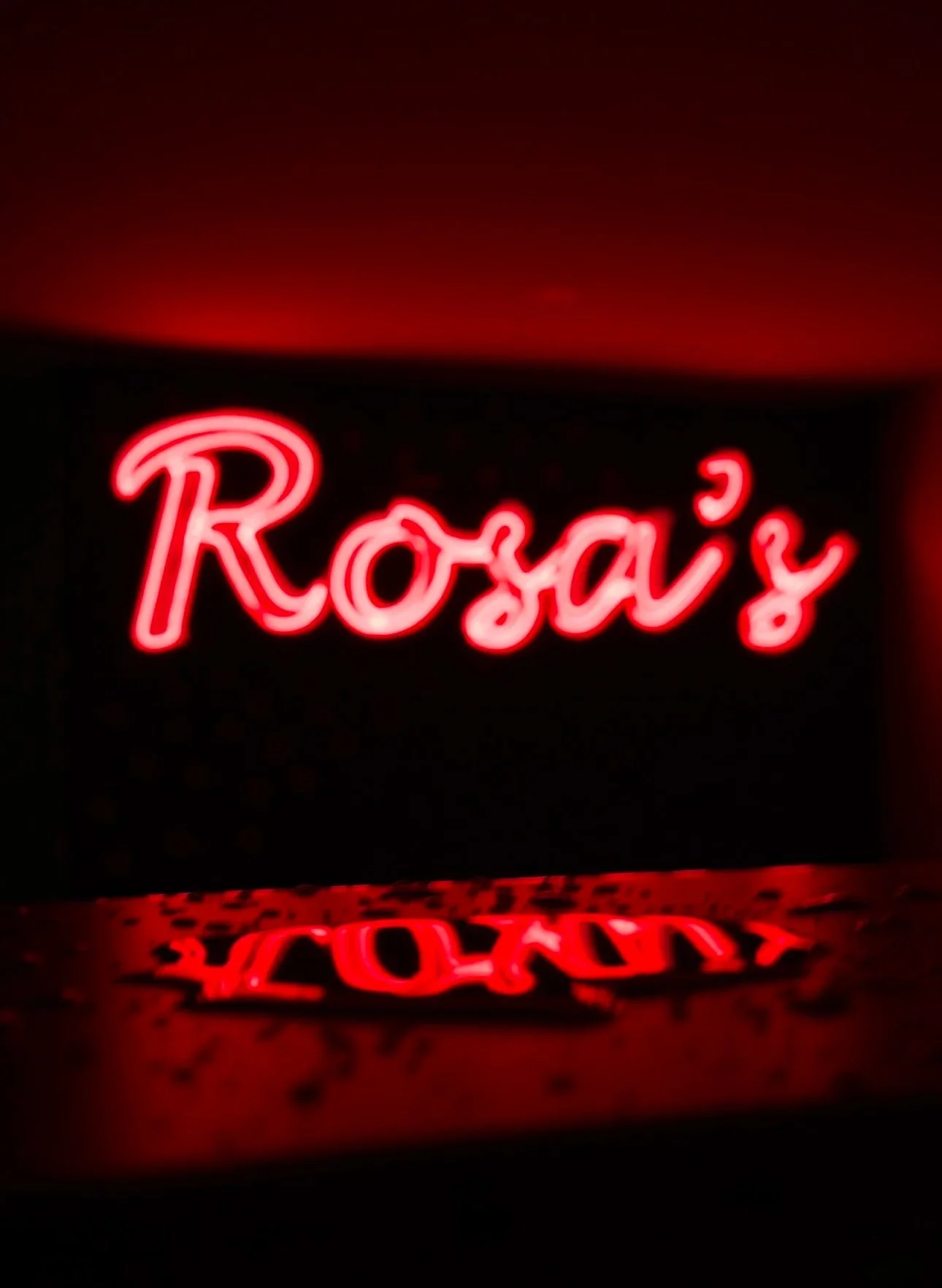 Red neon sign that reads 'Roxy' with its reflection on a shiny surface below.