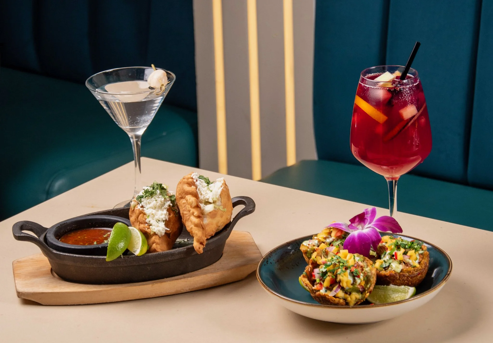 A table with two cocktails, two Mexican dishes, and lime wedges, set in a restaurant. One cocktail is a clear martini with an olive, and the other is a red drink with fruit slices and a straw. The dishes include two fried items topped with cheese and