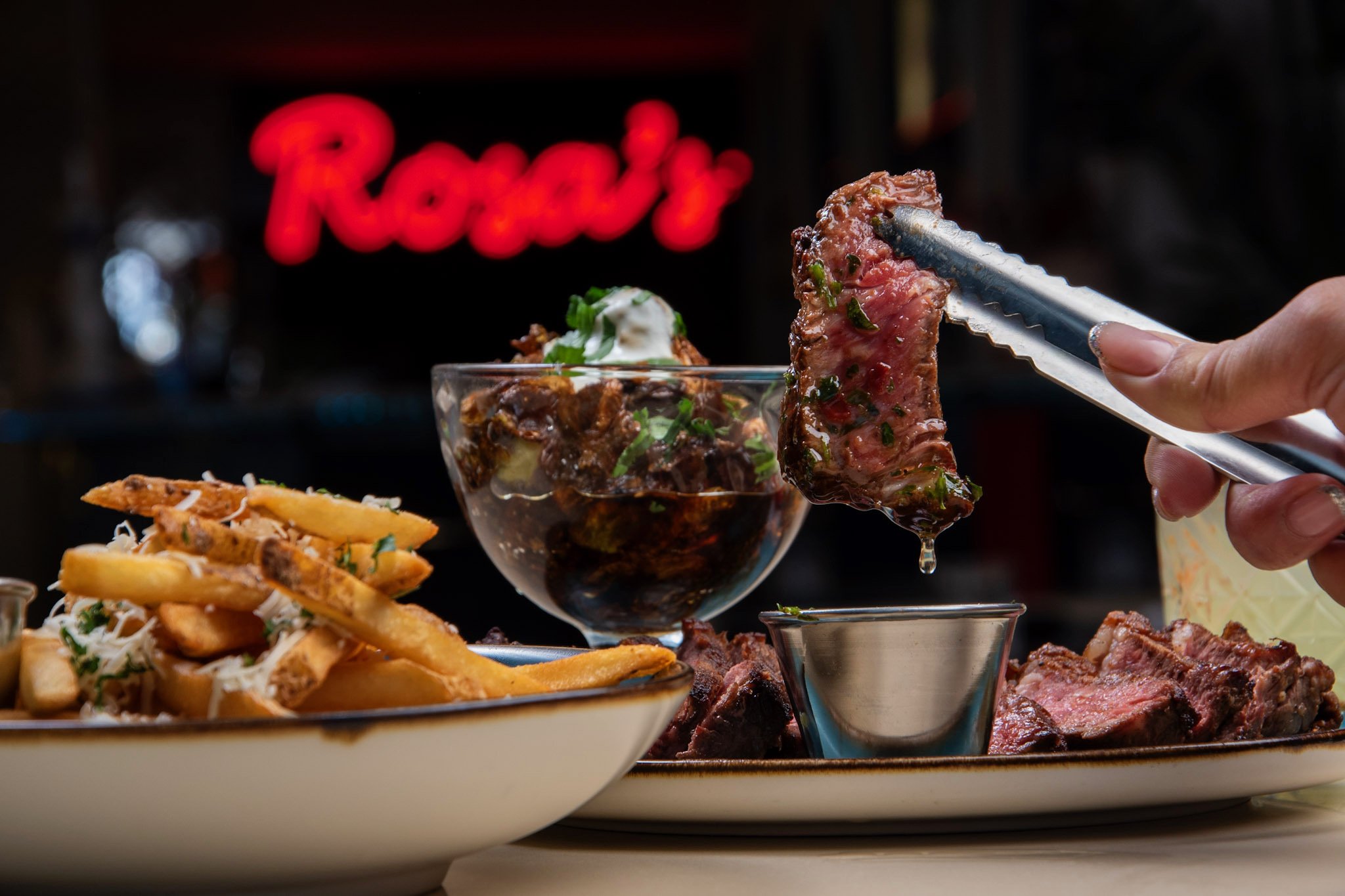 Best steak in Toronto - A plate of French fries with shredded cheese and herbs, a bowl of beef stew topped with sour cream and herbs, and a person holding a tongs with a piece of cooked steak