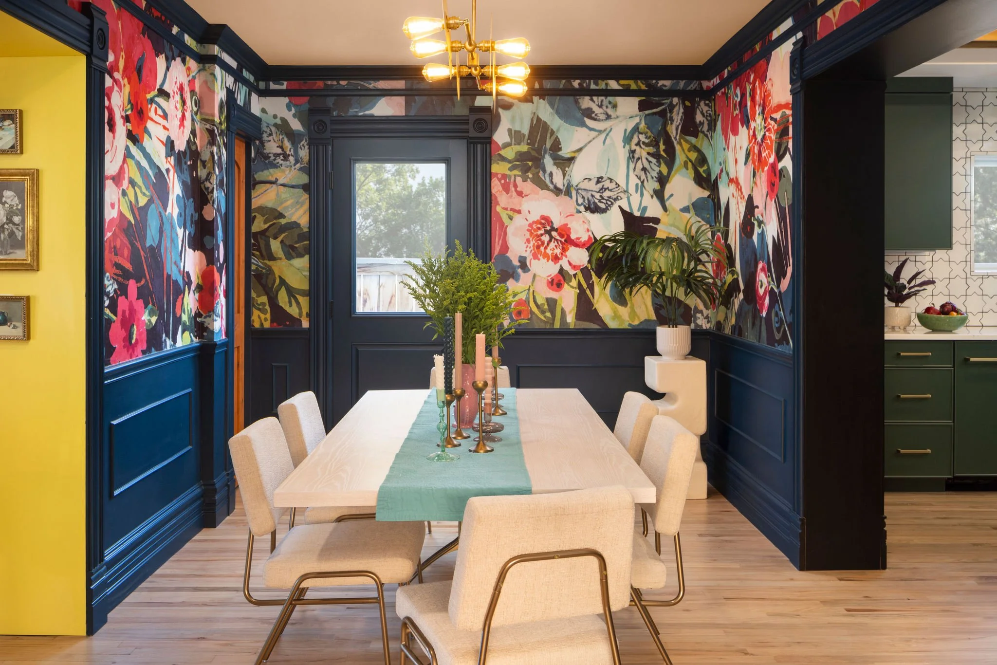 A dining room with a light wood table, beige chairs, a green table runner, and colorful floral mural walls. The table is decorated with candles and a plant centerpiece. There is a window in the background and a yellow wall on the left.