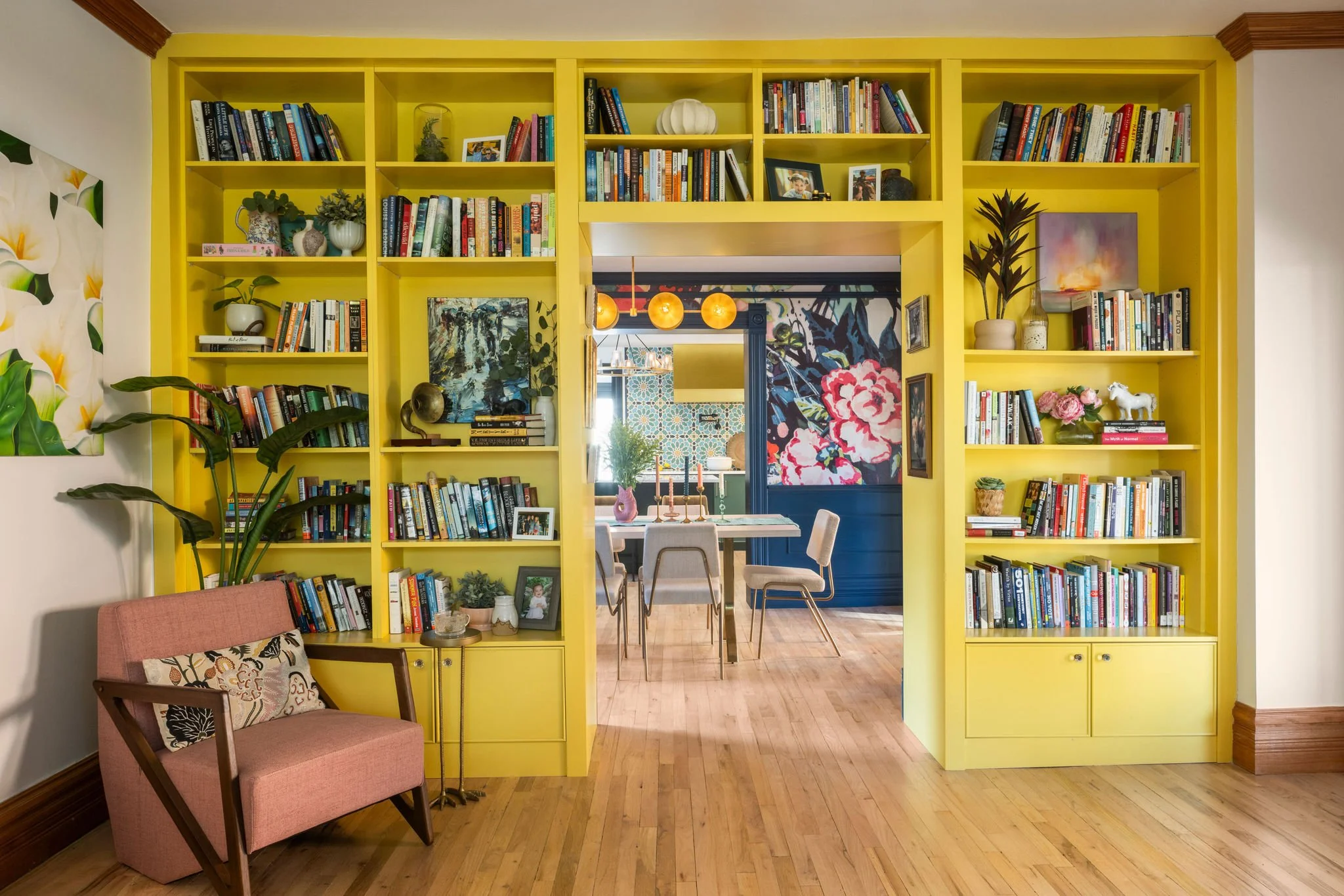 Bright yellow built-in bookshelves filled with books and decorative items frame a doorway leading to a dining area with floral wallpaper. A pink armchair with patterned pillow and a large green plant are on the left side of the room. The space has hardwood floors and crown molding.