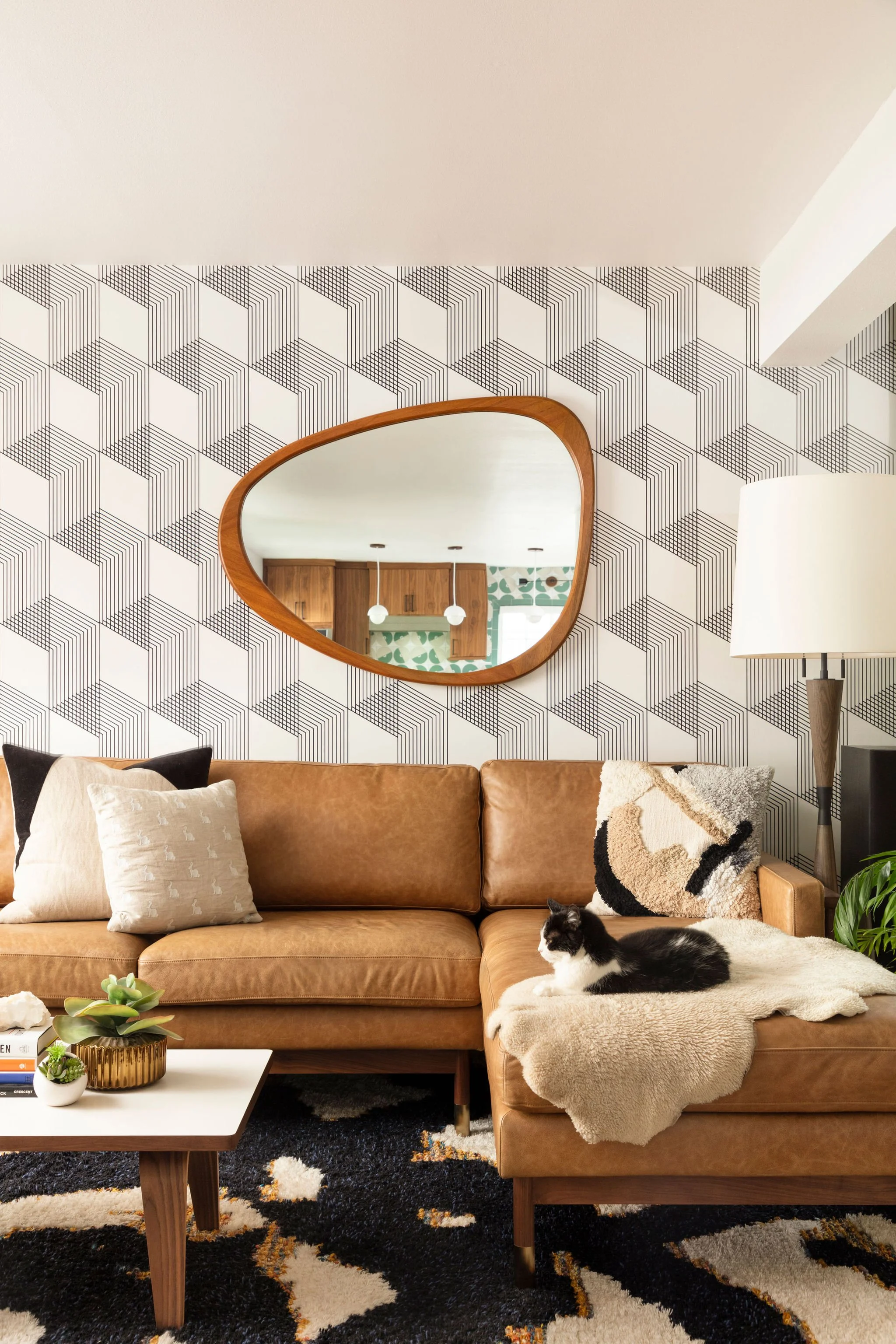 Living room with a brown leather sofa, decorative pillows, a white and black cat, a table with potted plants, a patterned black and white rug, a tall white lamp, and a geometric-patterned wallpaper with a wooden-framed mirror reflecting a kitchen with wooden cabinets and white pendant lights.