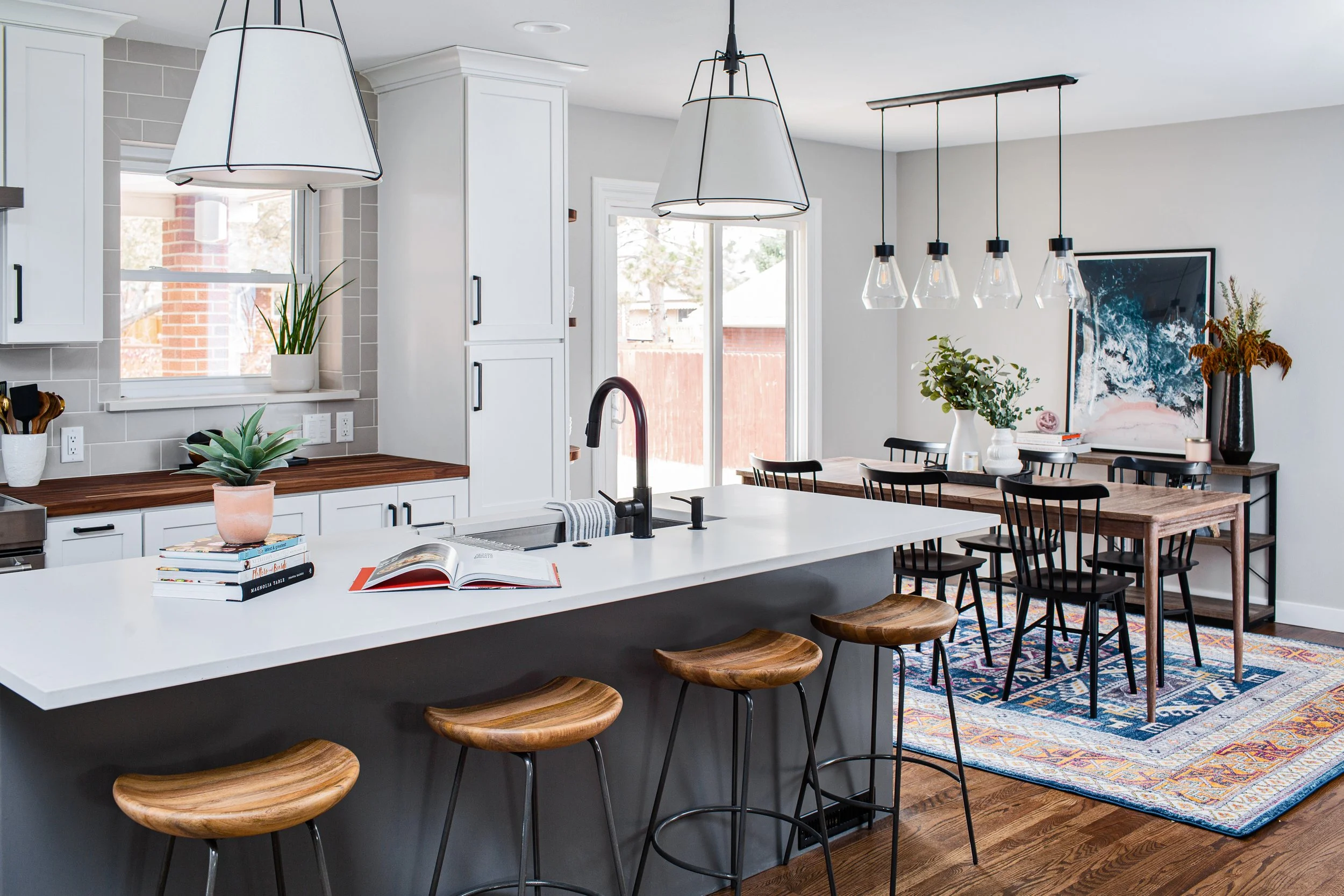 Modern kitchen and dining area with white cabinets, a white kitchen island with wooden stools, a dining table with black chairs, colorful area rug, pendant lights, and decorative plants.
