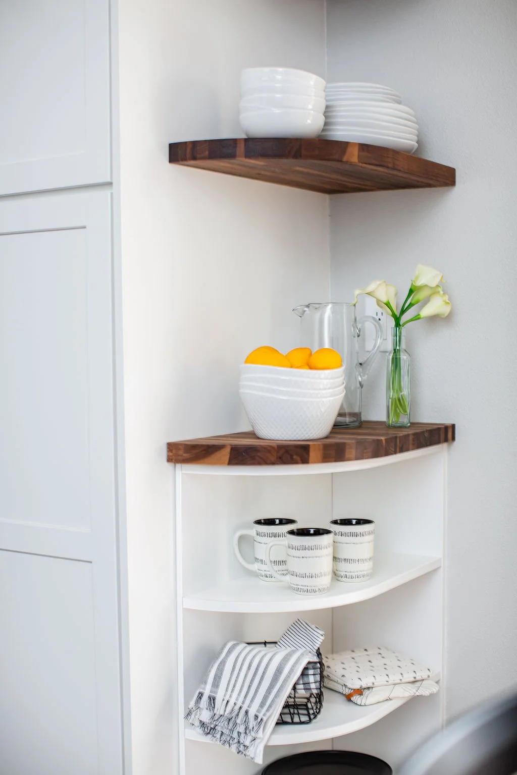 Corner kitchen shelf with white dishes on top, a bowl of lemons, a glass pitcher, a vase of white calla lilies, black and white striped mugs, and kitchen towels.