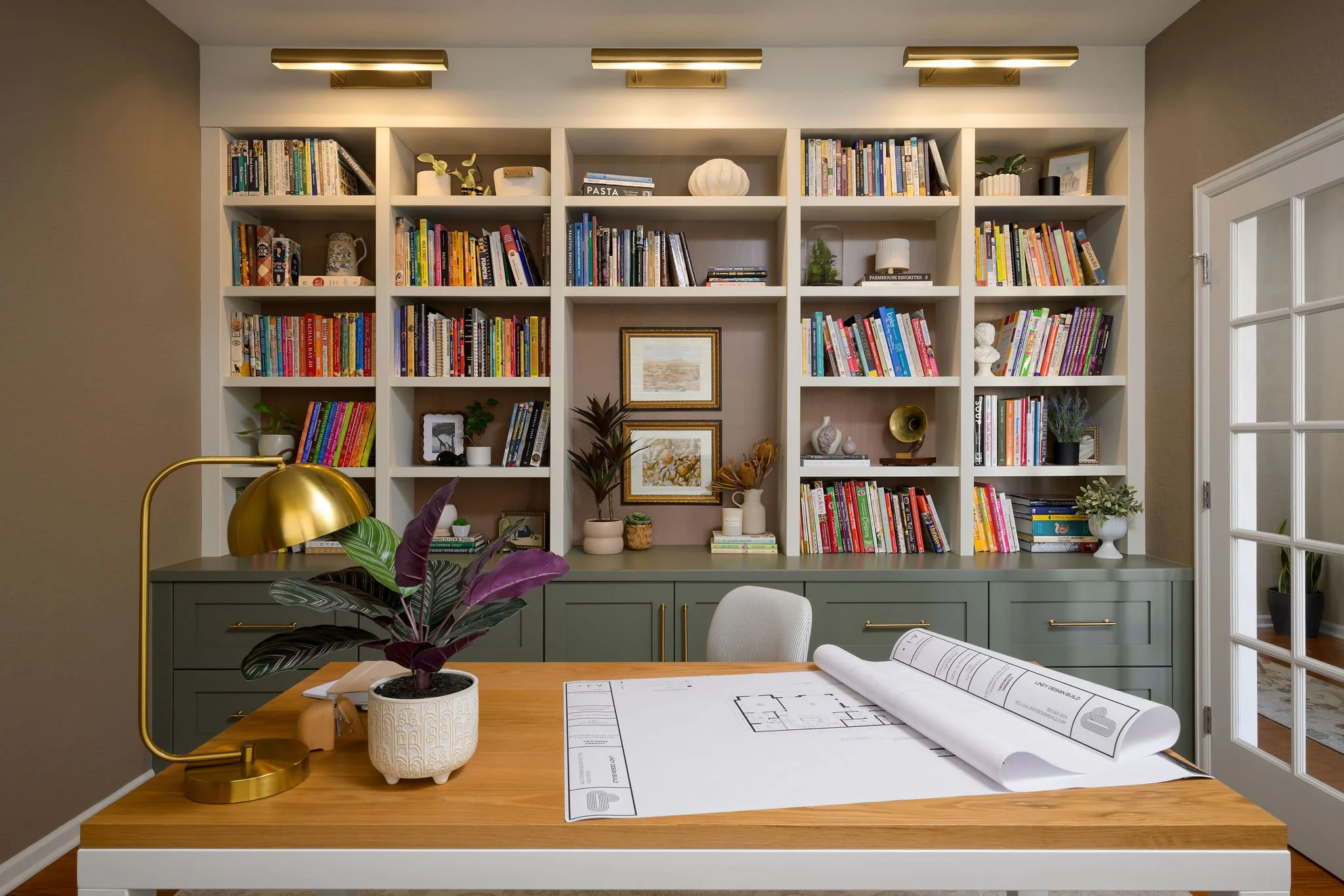 Interior of a home office with a wooden desk, a potted plant, a gold desk lamp, and a set of architectural blueprints. In the background, white built-in bookshelves filled with colorful books, decorative items, and framed pictures, and a gray cabinet below. A glass door with white trim is on the right side.