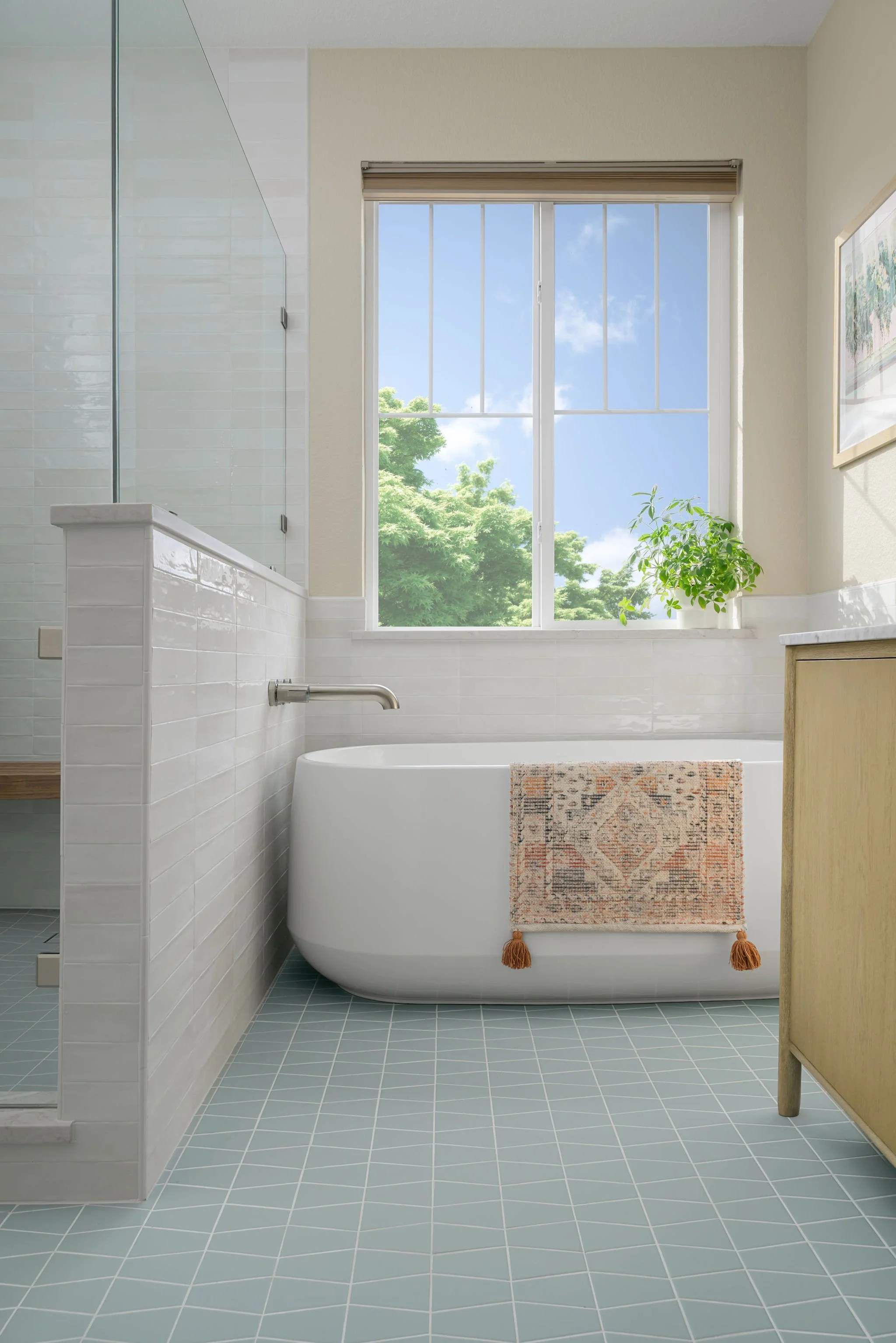 Modern bathroom with a bathtub near a window showing green trees and a blue sky, with a small potted green plant on the windowsill and a decorative towel hanging on the bathtub.