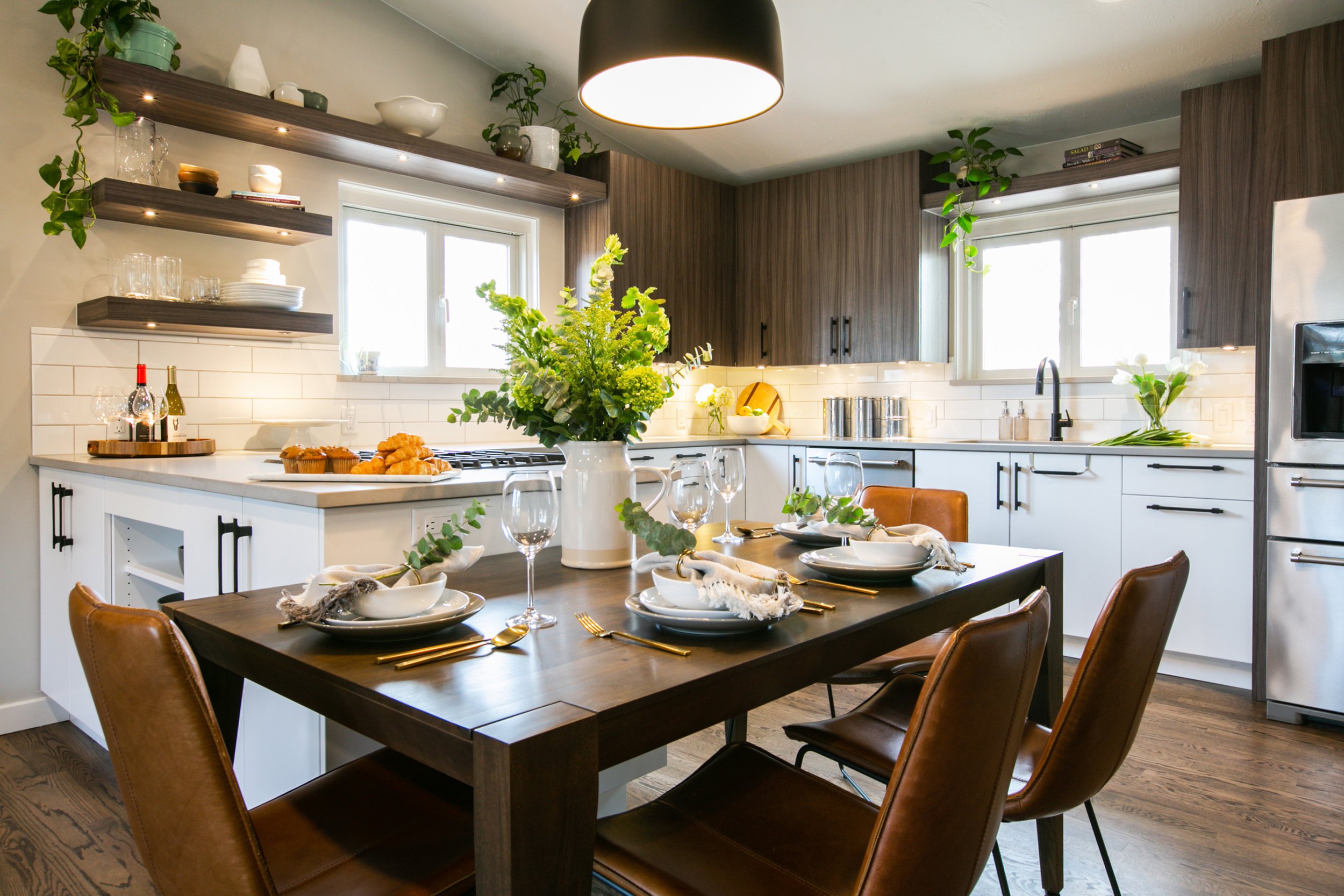 A modern kitchen and dining area with a dark wood table set for four, with white plates, bowls, gold utensils, and napkins. A large white pitcher with green foliage and flowers serves as a centerpiece. The kitchen has white cabinets, a white tile backsplash, and dark wood upper cabinets. There are two windows above the kitchen sink, which has a black faucet. Open shelves hold glassware, dishes, and plants. A black pendant light hangs over the table. The floor is wood, and the overall decor is contemporary and cozy.