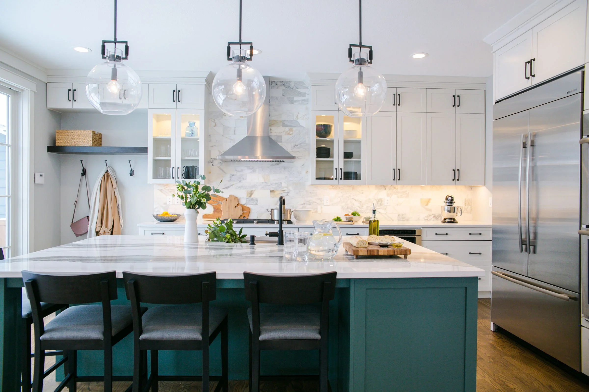 Modern white kitchen with marble backsplash, stainless steel appliances, hanging globe pendant lights, and a teal island with black chairs.