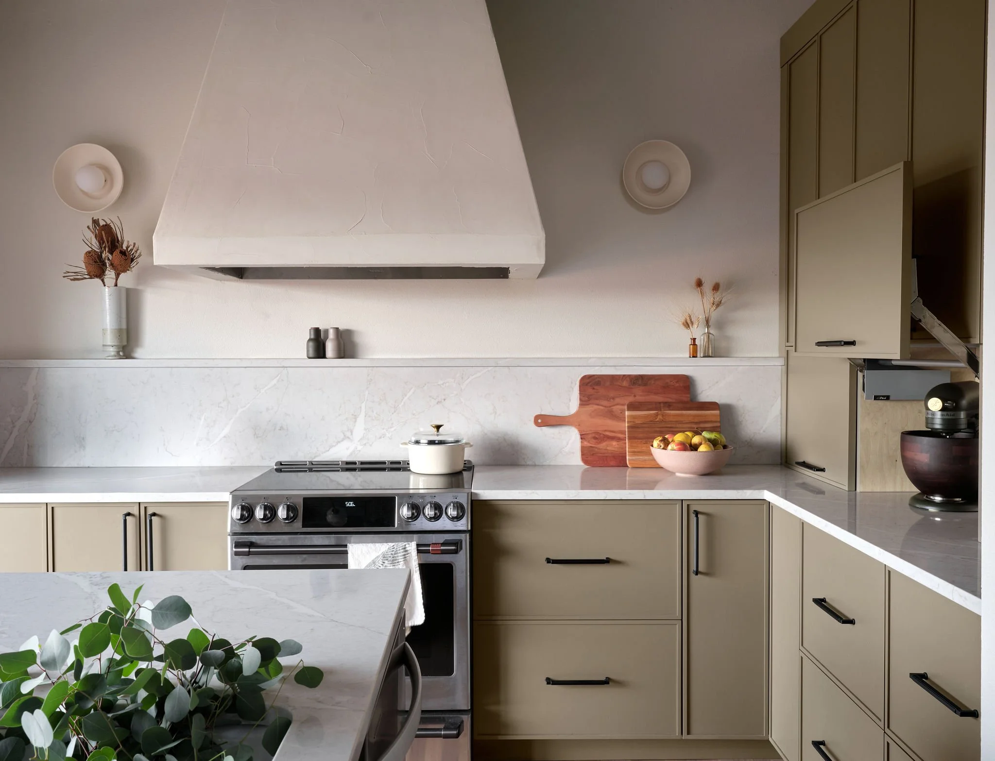 A modern kitchen with beige cabinets, a white marble countertop, and a stove with a white pot. Decor includes dried flowers and bowls of apples, with a large white range hood and neutral wall-mounted lights.