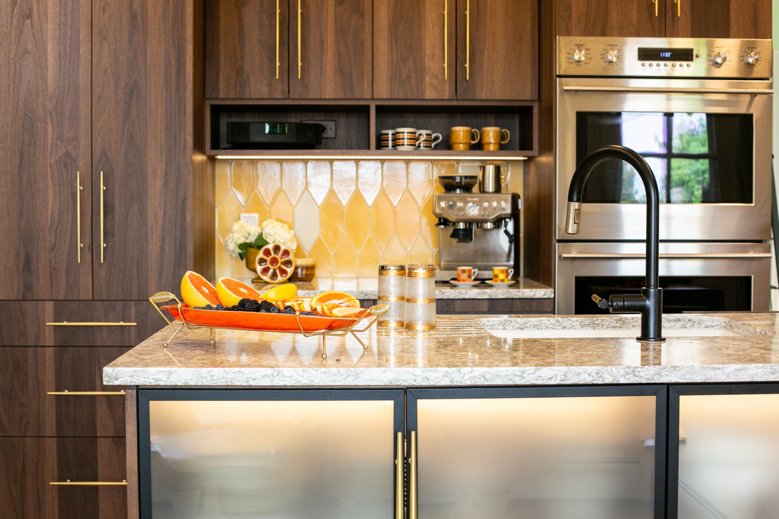 Modern kitchen with wooden cabinets, stainless steel oven, black faucet, marble countertop, and a fruit platter with oranges, blackberries, and strawberries.