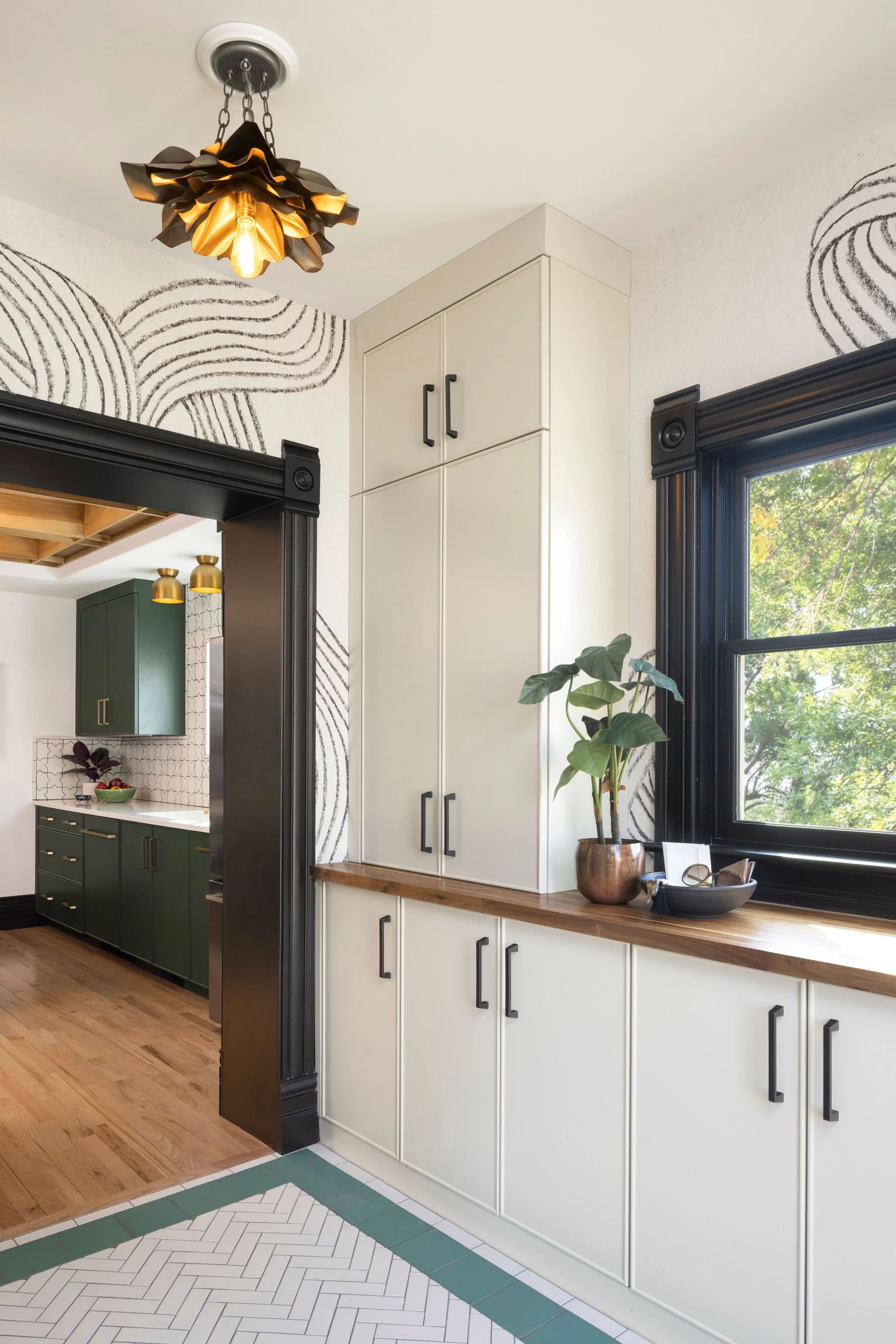 Interior view of a kitchen with black trim around window and doorway, white cabinets with black handles, and a wooden countertop. There is a potted plant on the countertop near a window, and a darker green kitchen area visible through a doorway. The ceiling features a decorative black and gold light fixture, and the wall has black line art patterns.