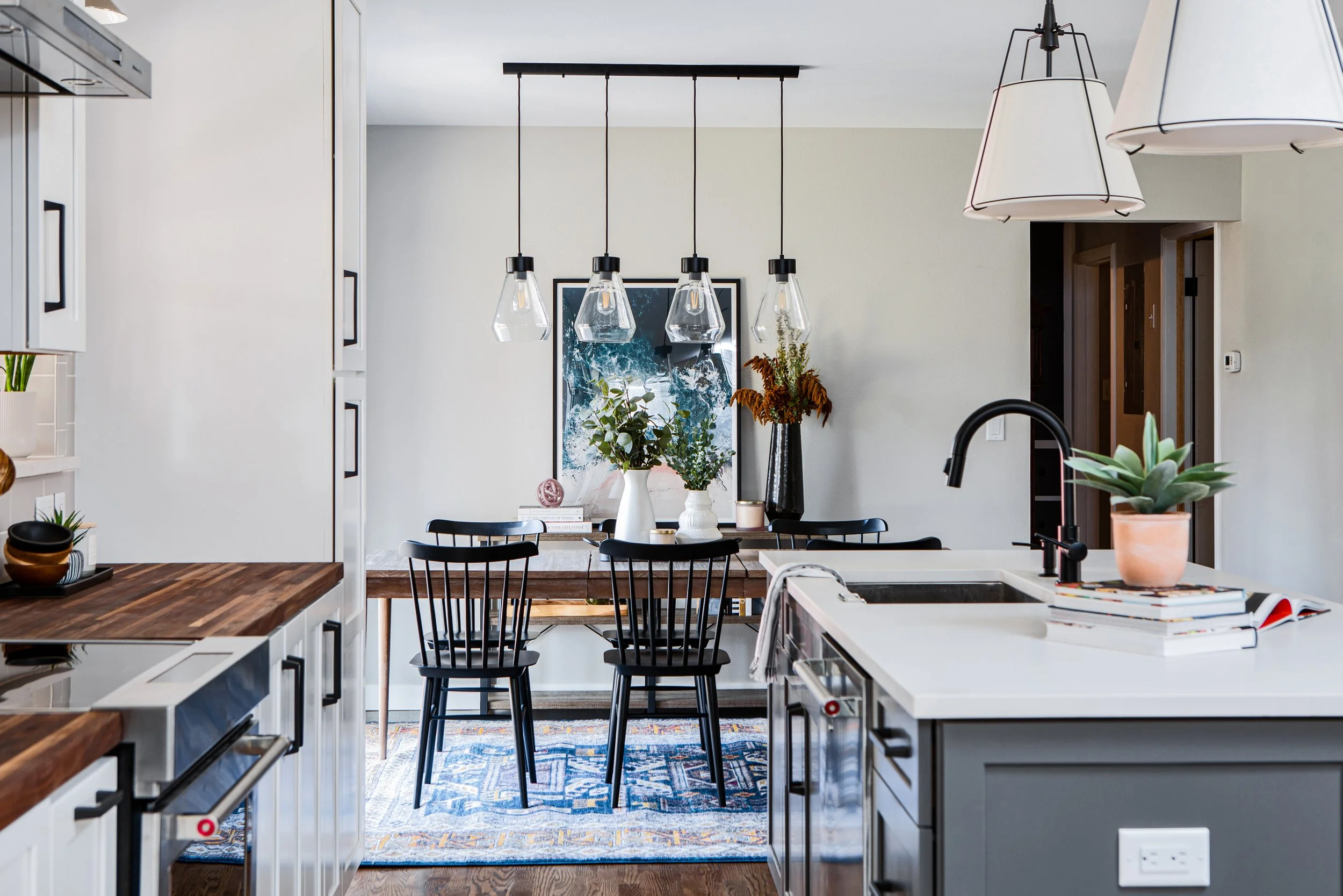 Modern kitchen with a white island counter, black faucet, and potted succulent plant, with a dining area featuring six black chairs, vases with plants, and hanging pendant lights.