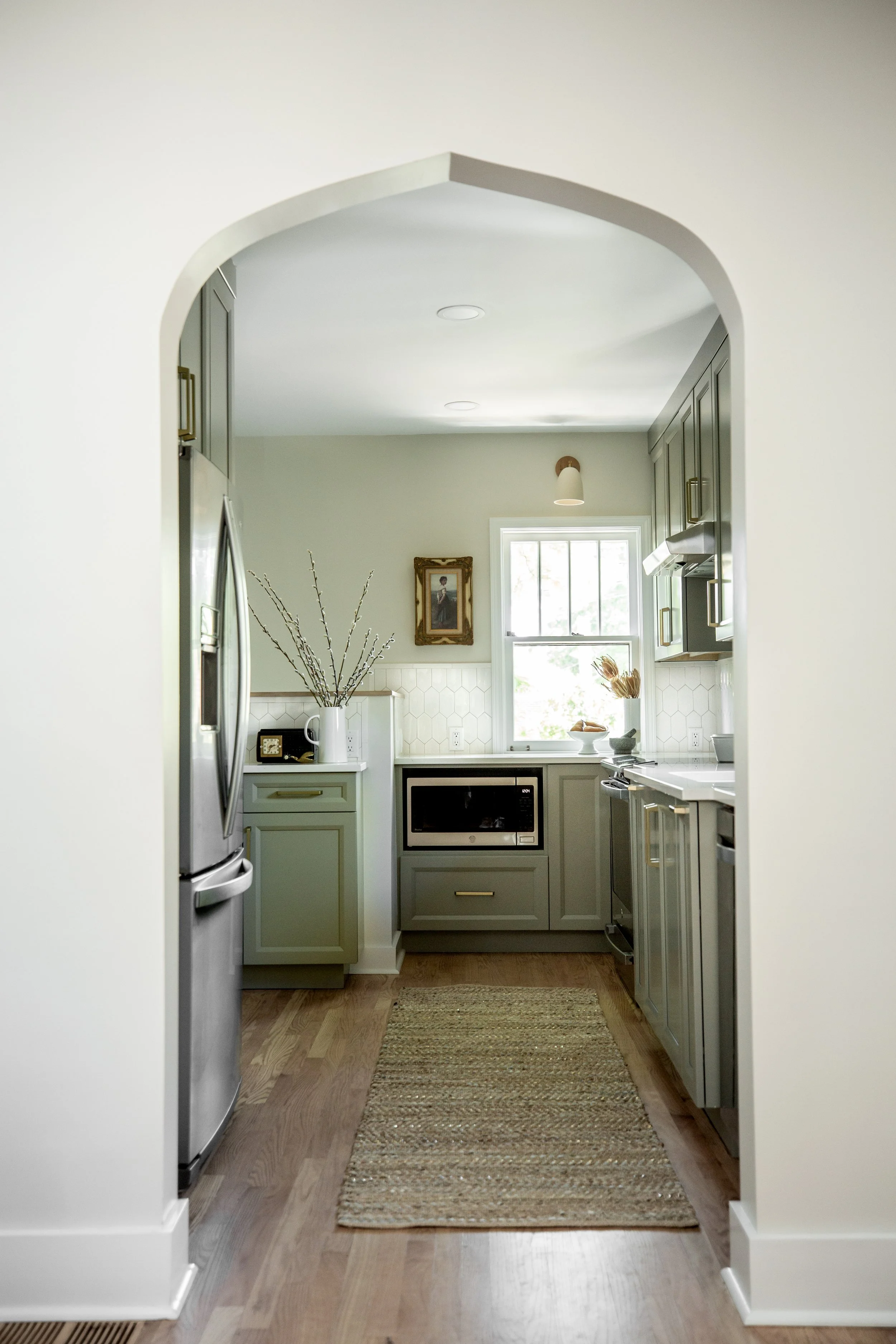 A modern kitchen with grey cabinets and wood flooring, viewed through an arched doorway. The kitchen has a window with natural light, a microwave, and decorative items on the countertop.