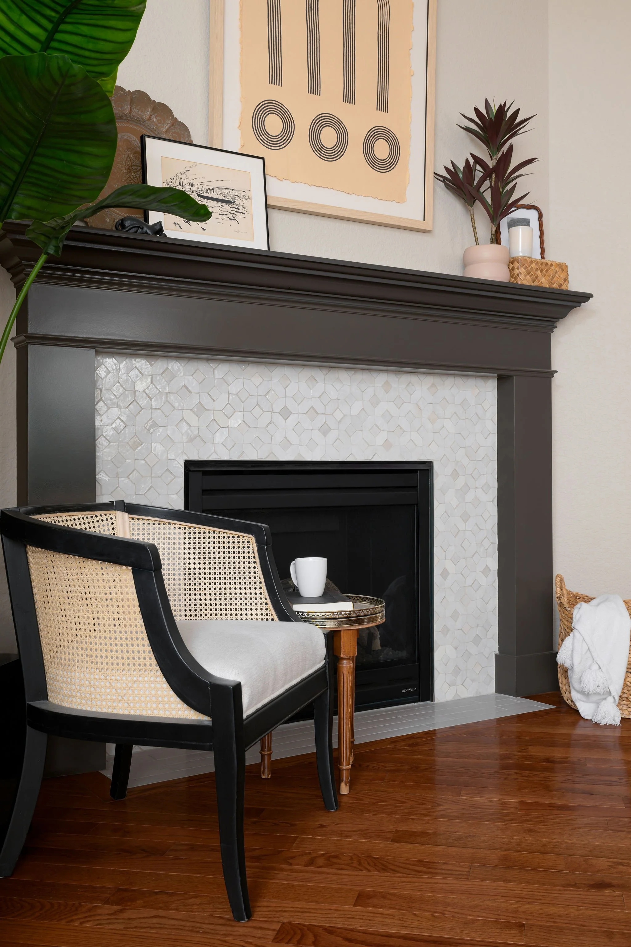 Living room with a dark gray fireplace, beige tile surround, a black-and-white framed drawing on mantel, and abstract artwork above. A black chair with light cushion, small wooden side table with white mug, potted plant, and wicker basket with blanket on the hardwood floor.