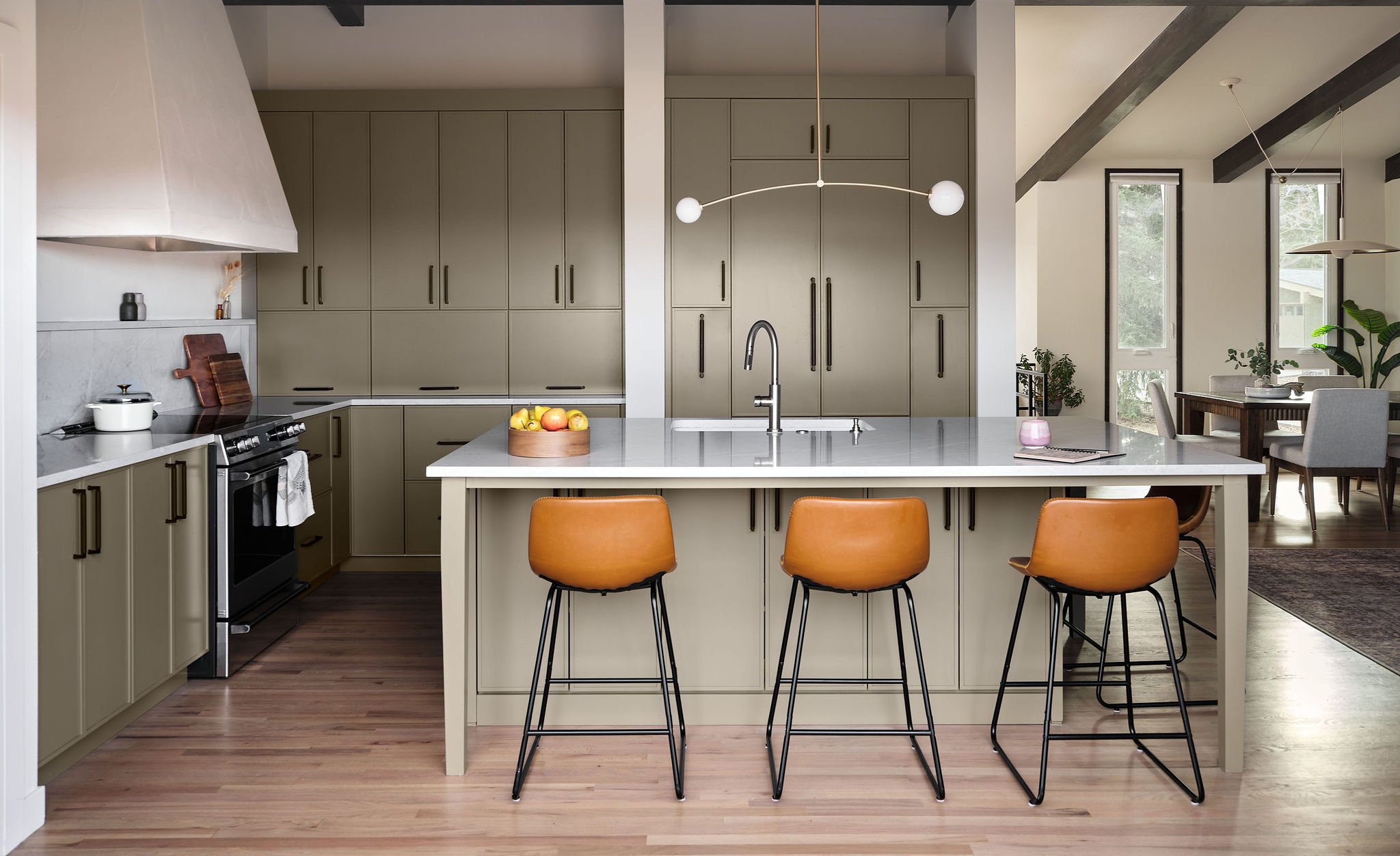 Modern kitchen with green cabinets, white island with three tan barstools, fruit bowl on island, black stove, and open dining area with large windows and greenery outside.