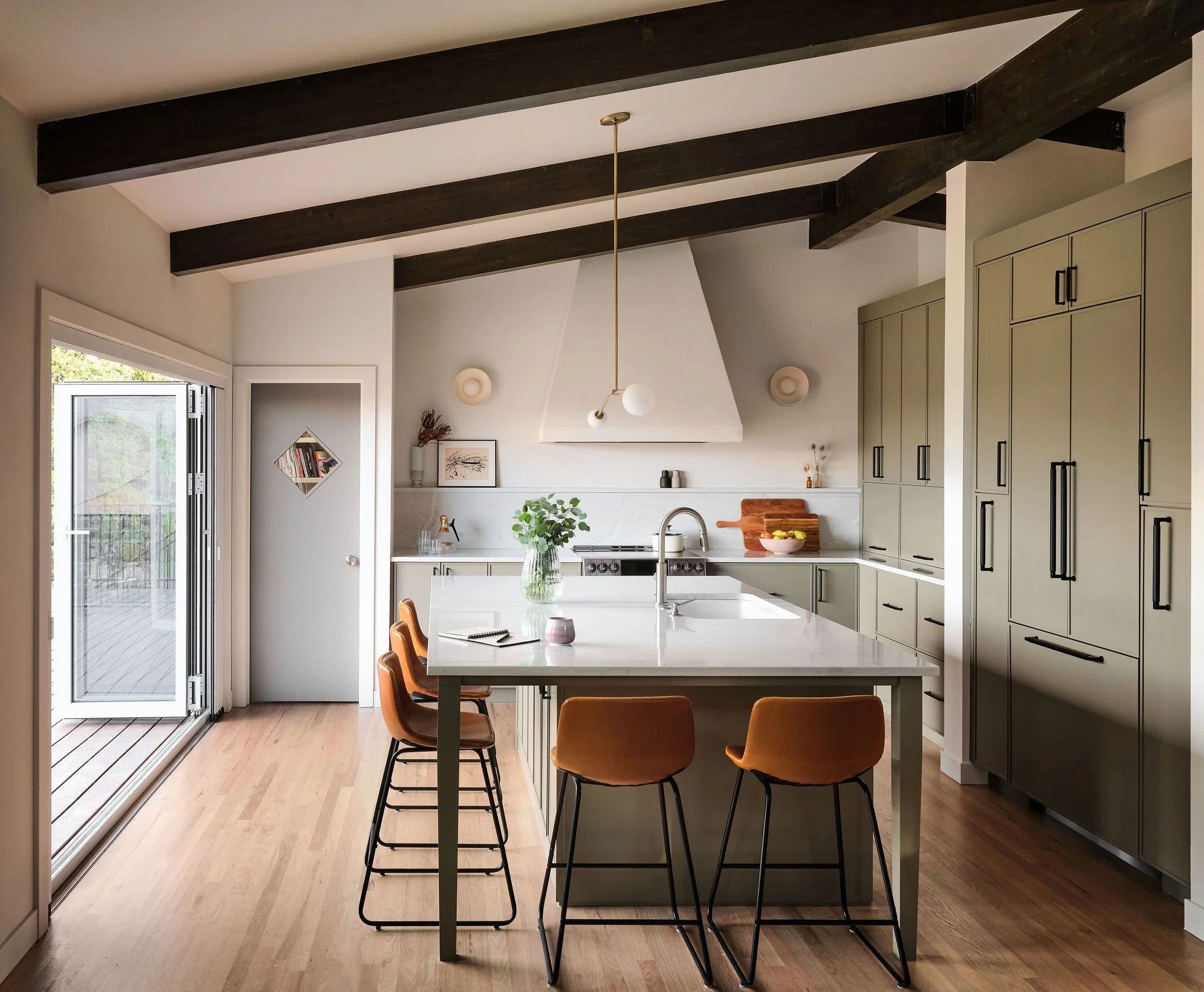 Modern kitchen with light green cabinetry, a central island with a marble countertop, five orange barstools, a sliding glass door, a fireplace with a white chimney, and decorative wall accents.