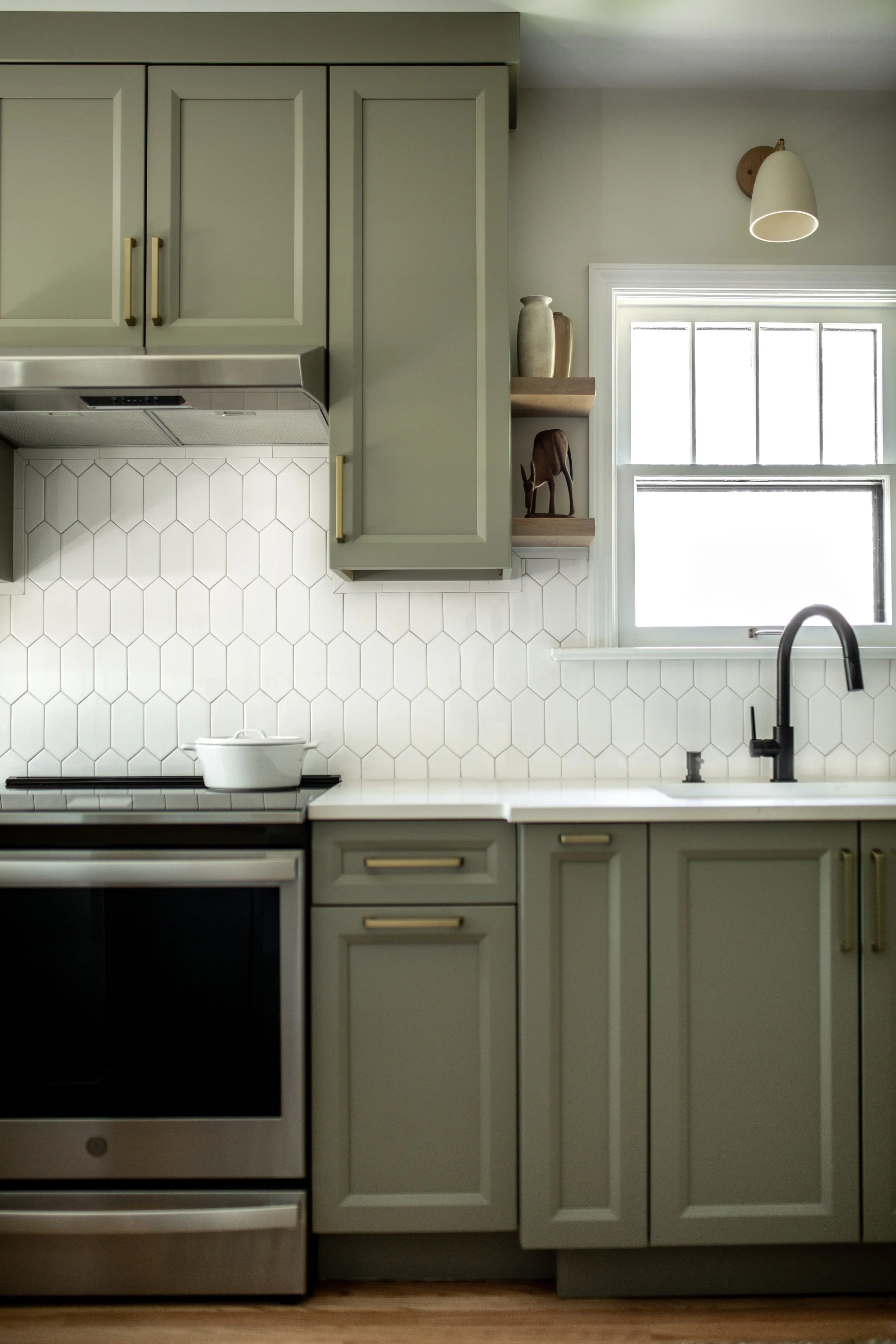 Green kitchen cabinets with gold handles, white hexagon tile backsplash, black faucet, black stove, and a window with a white frame and frosted glass. Small shelves hold vases and a decorative horse figure.