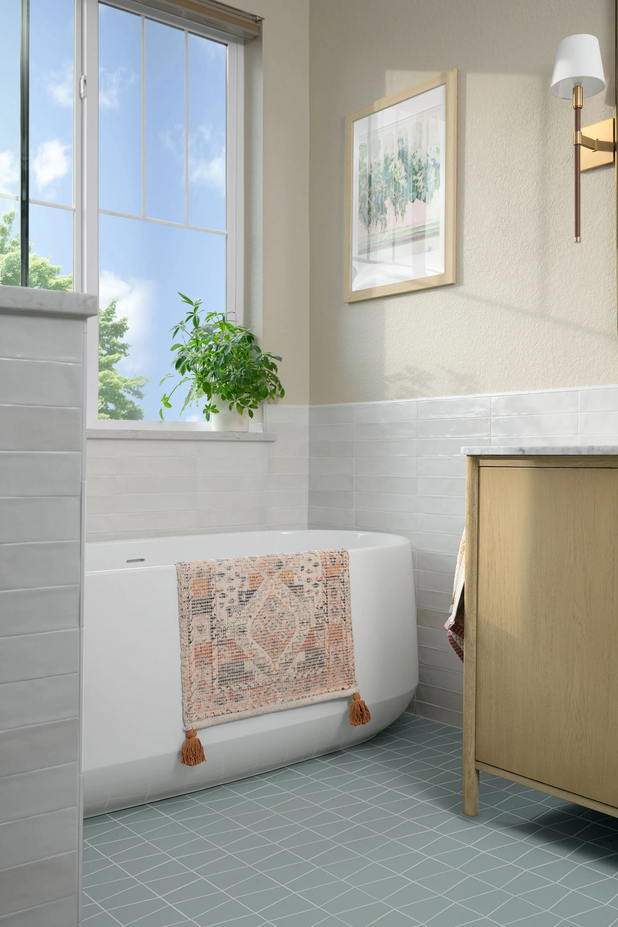 A bathroom with a large window, potted plant, bathtub with a patterned towel hanging over the edge, framed artwork, wall sconce light, and a wooden cabinet.