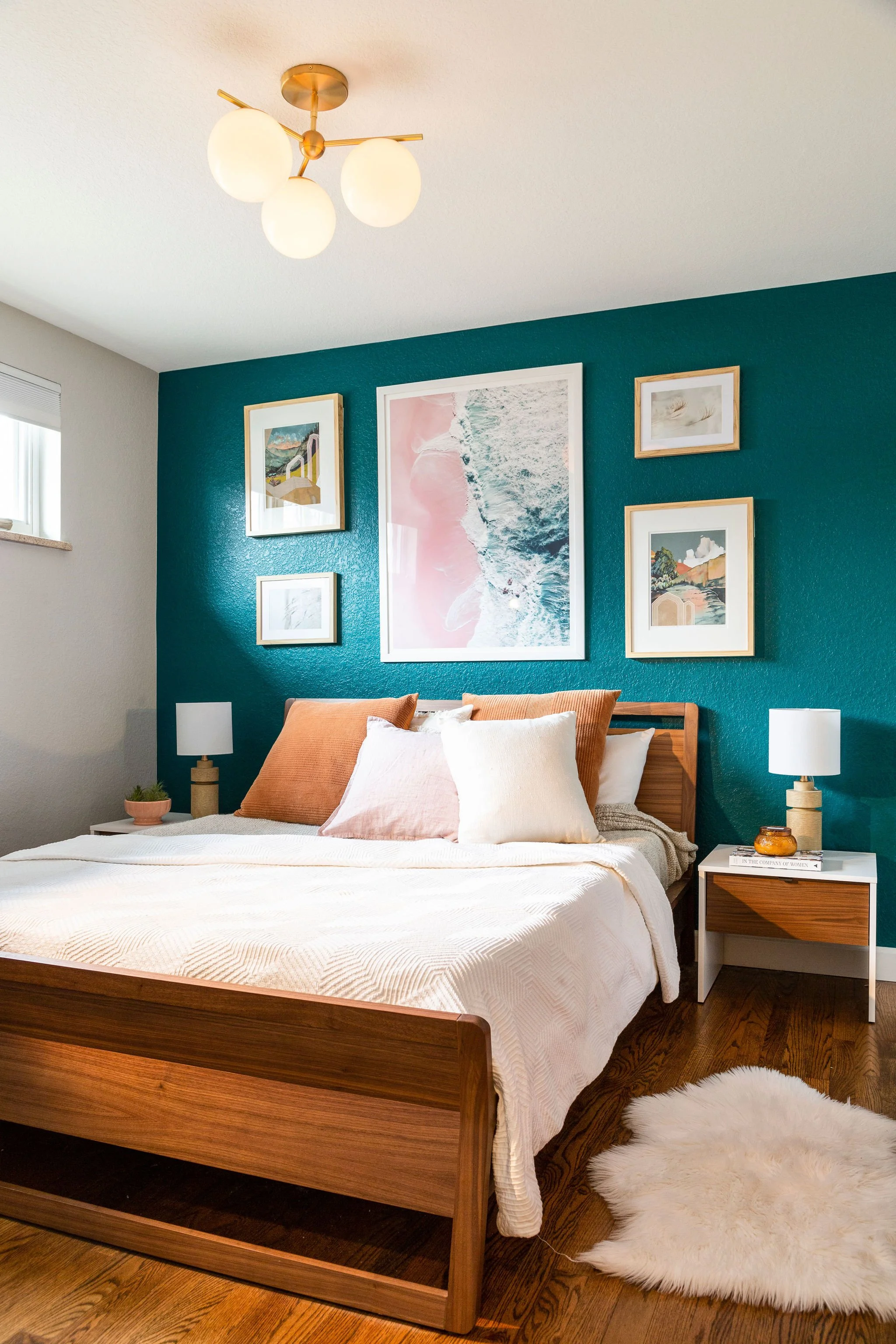 A bedroom with a wooden bed, white bedding, and decorative pillows, flanked by two white nightstands with lamps, a teal accent wall with framed artwork, hardwood floors, and a white shaggy rug.