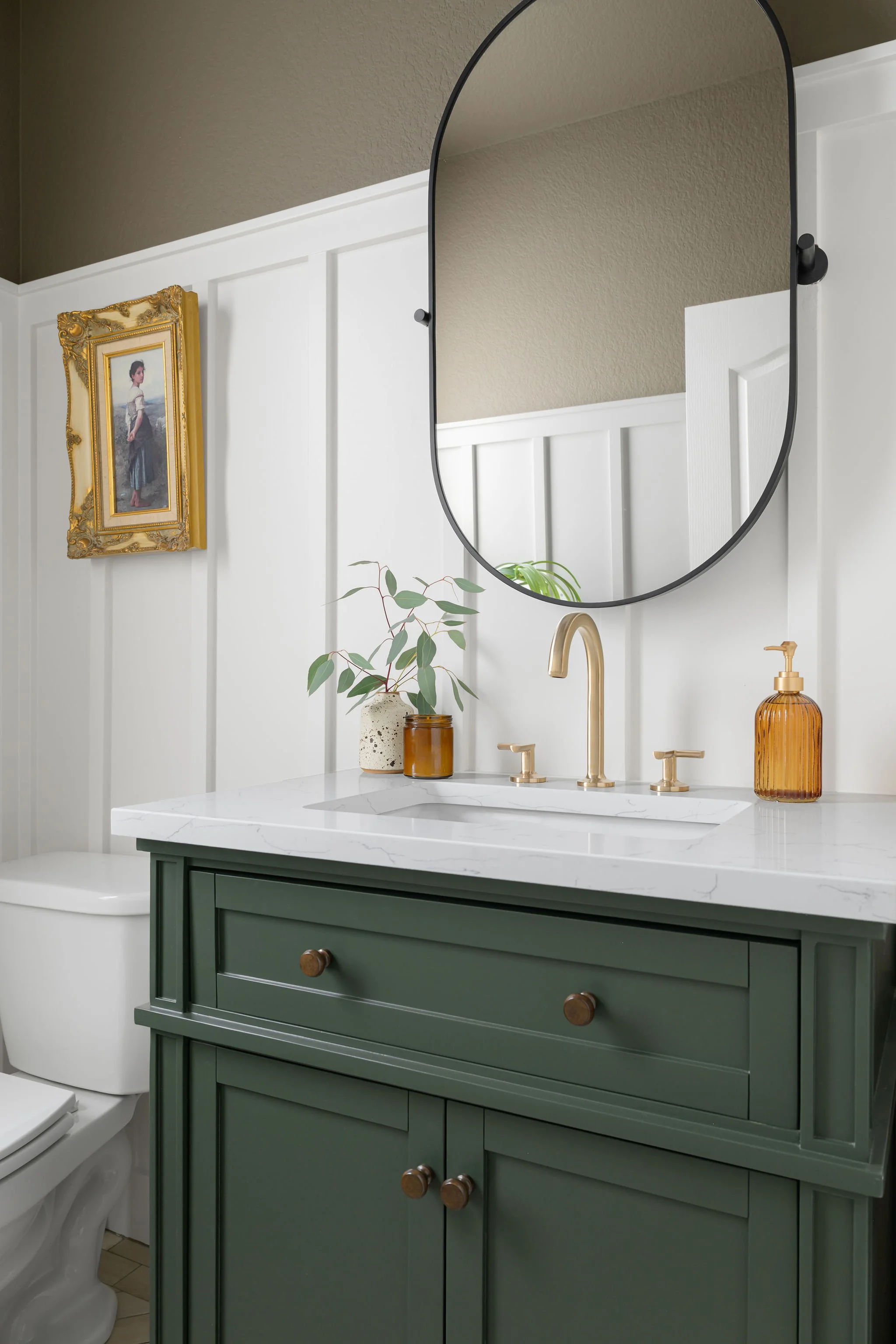 Bathroom with a green vanity, white marble countertop, gold faucet, oval mirror, amber soap dispenser, potted plant, framed artwork, and part of a toilet.