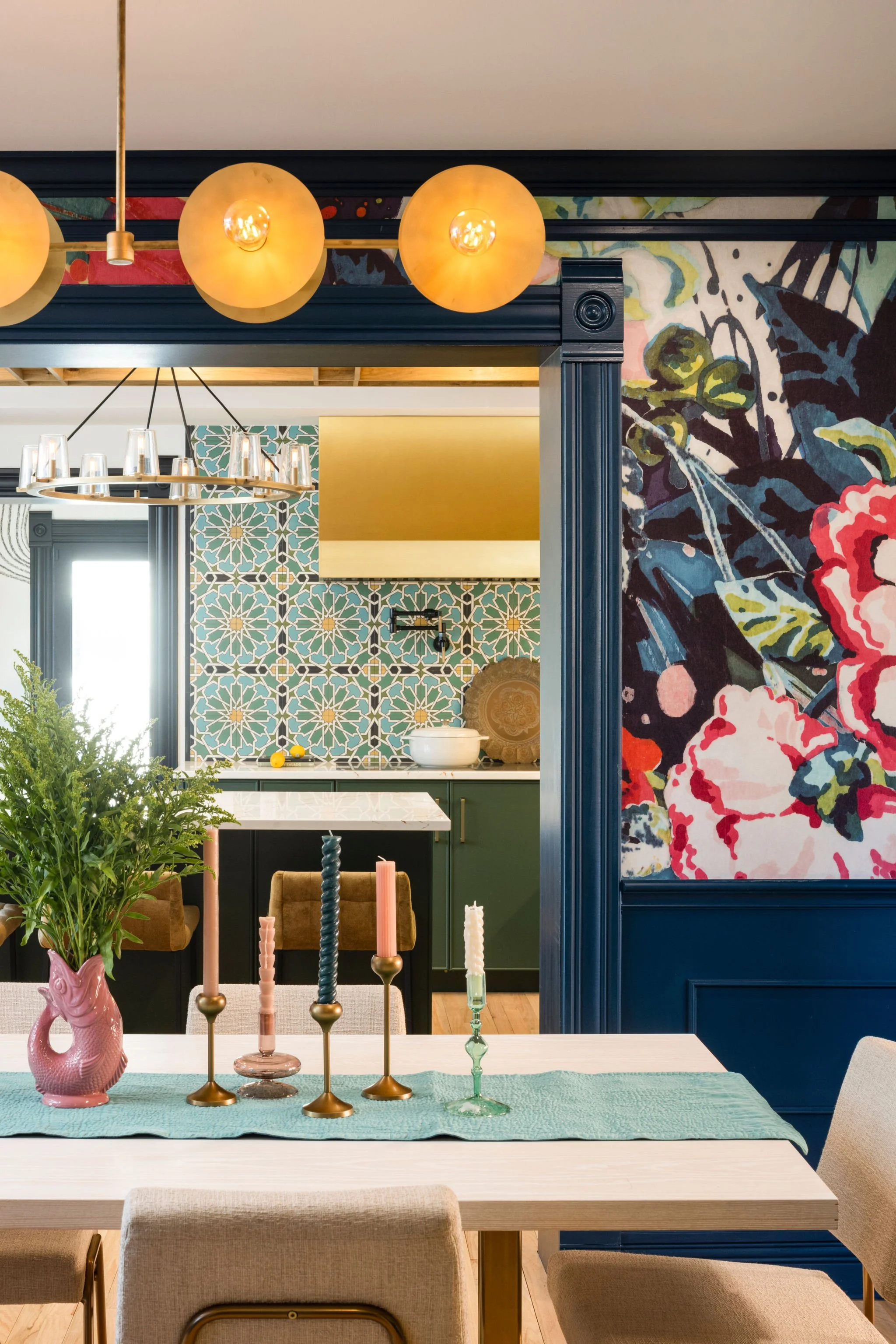 A dining room with a white table, beige chairs, and a blue table runner. On the table are five colorful candles in candlesticks and a pink vase with green plants. In the background, the kitchen has a vibrant tile backsplash, green cabinets, and a white countertop. A hanging light fixture and a wall with floral and geometric patterns are also visible.